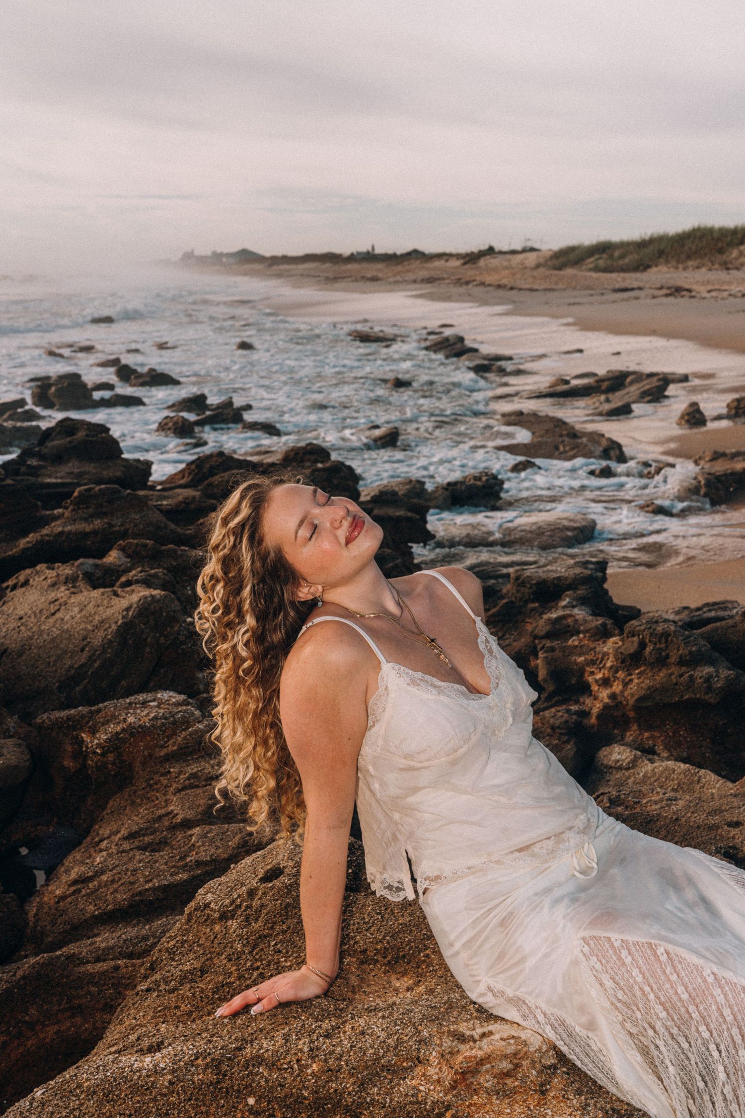 A woman in a white lace dress sits on rocky shoreline with eyes closed, waves and sandy beach in background.