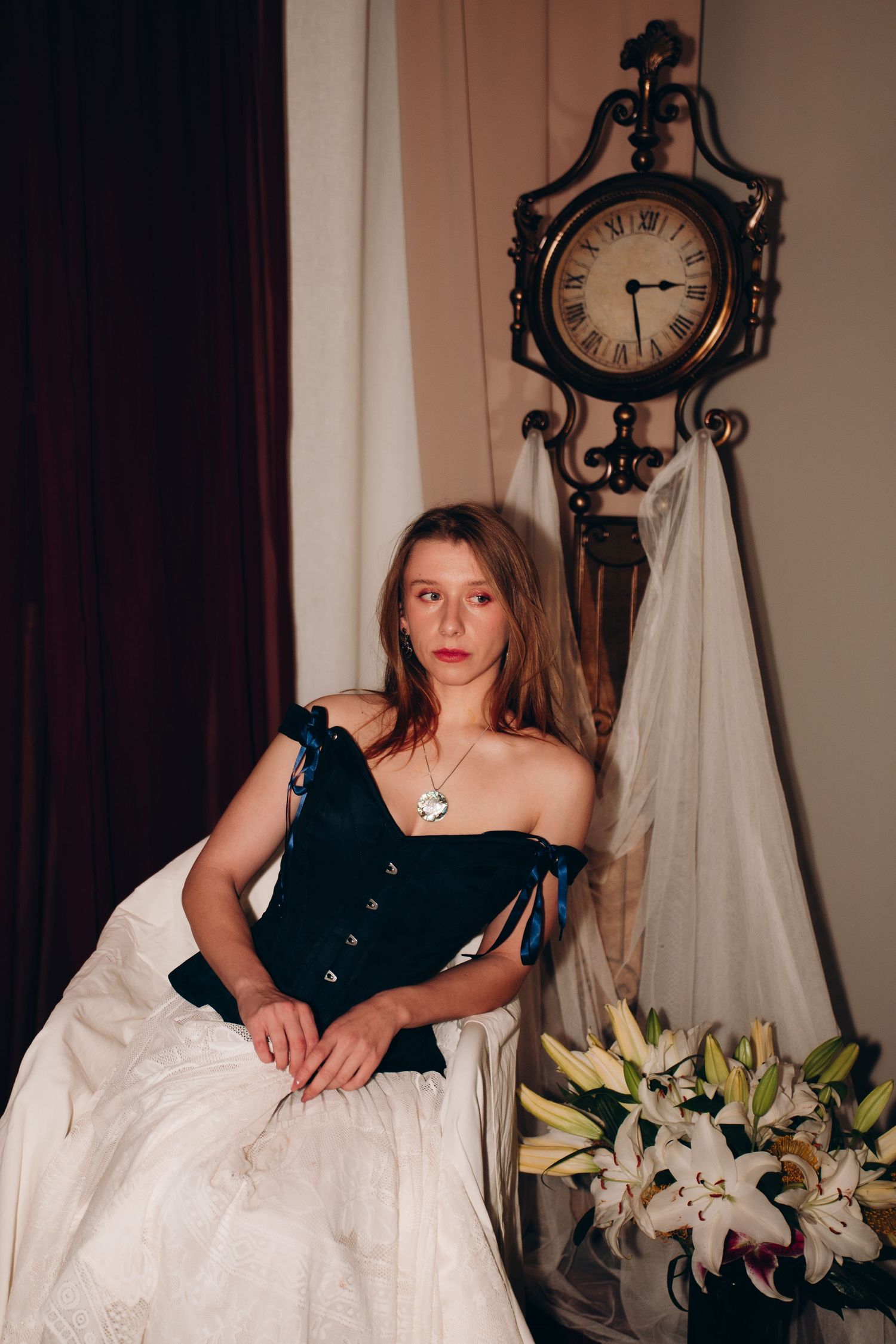 Young woman in dark velvet corset and white skirt poses near ornate vintage clock with white lilies nearby.