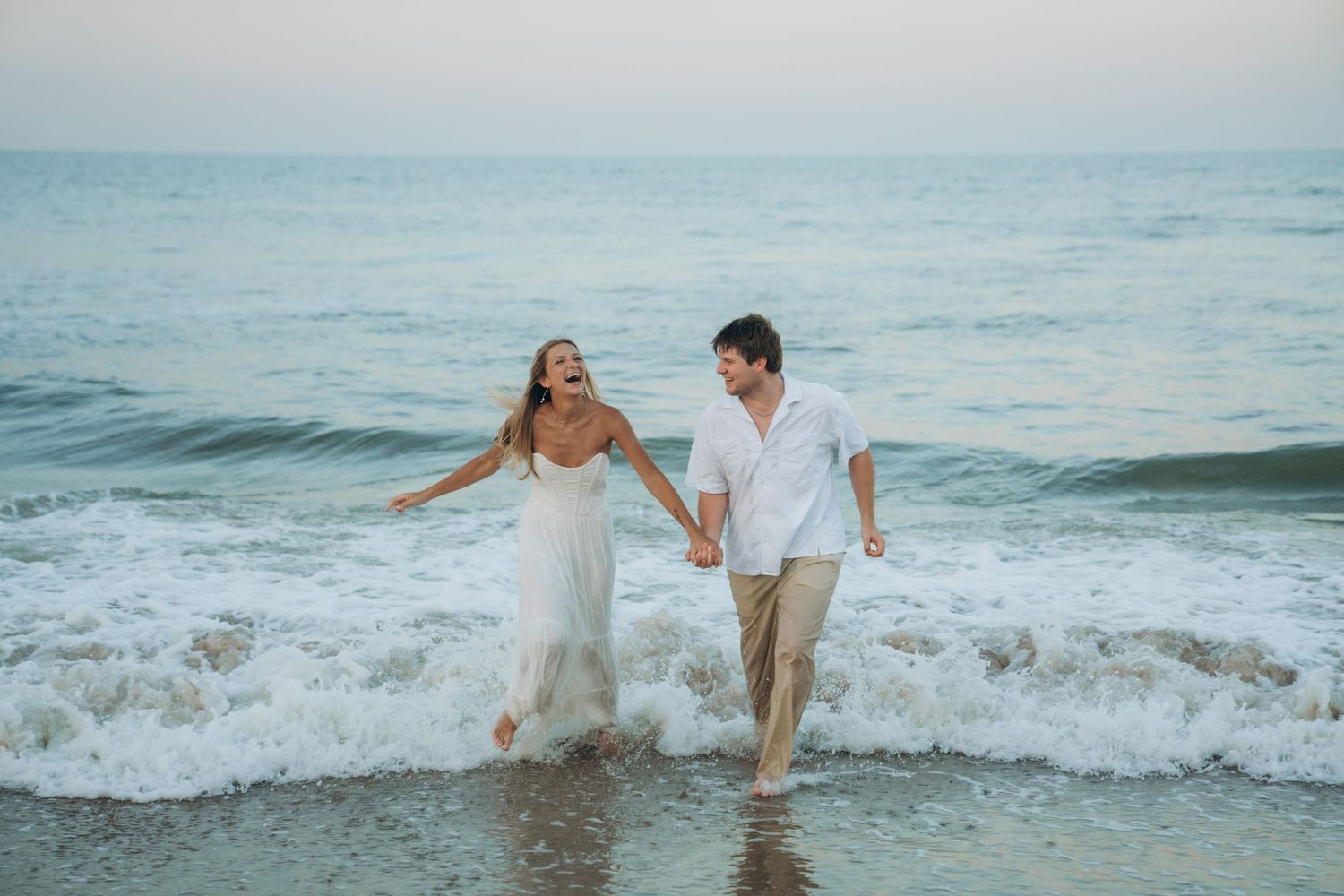 Couple splashing and playing in ocean waves wearing white outfits.