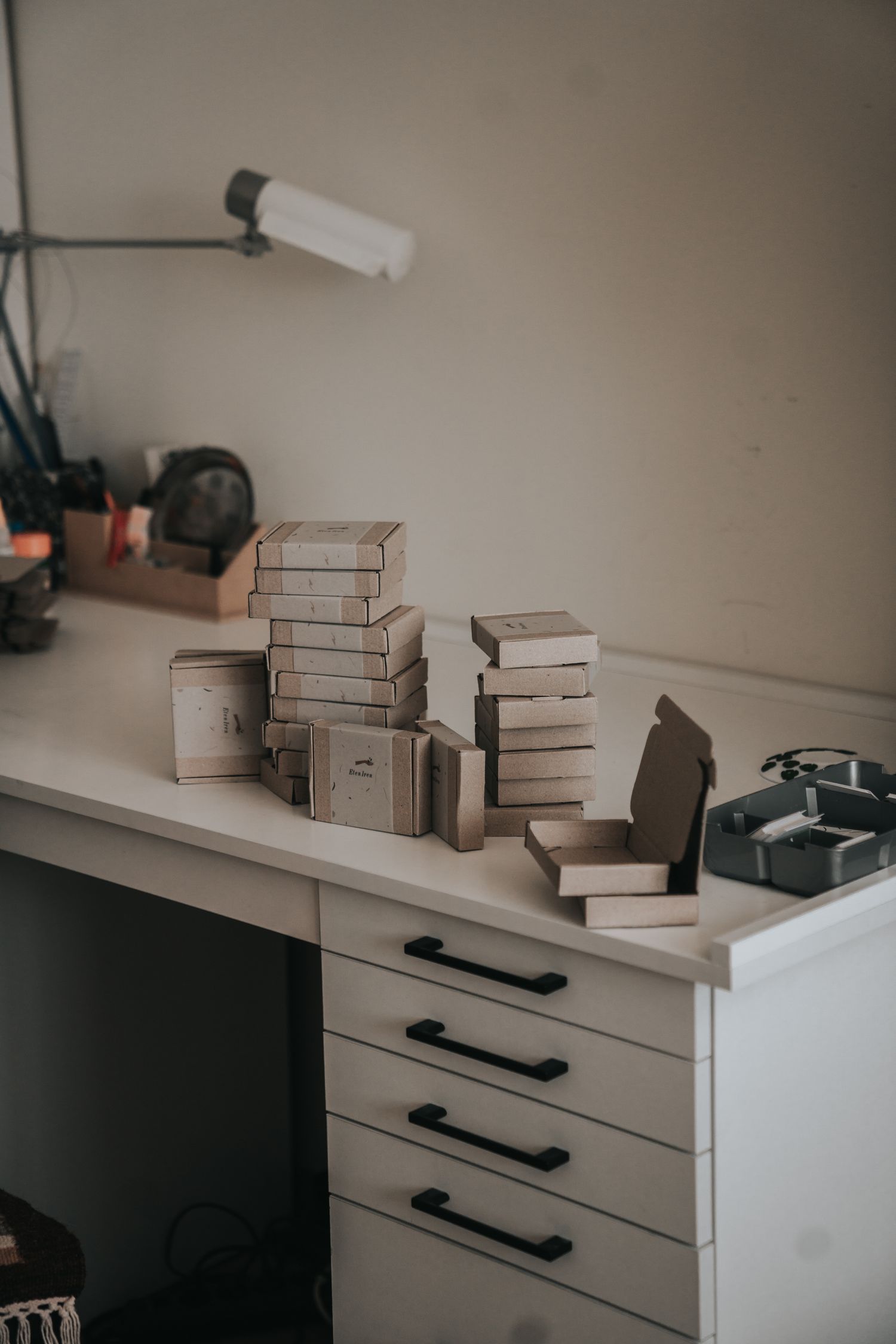 Stacks of small cardboard gift boxes arranged on a white drawer unit with a lamp in a craft studio.
