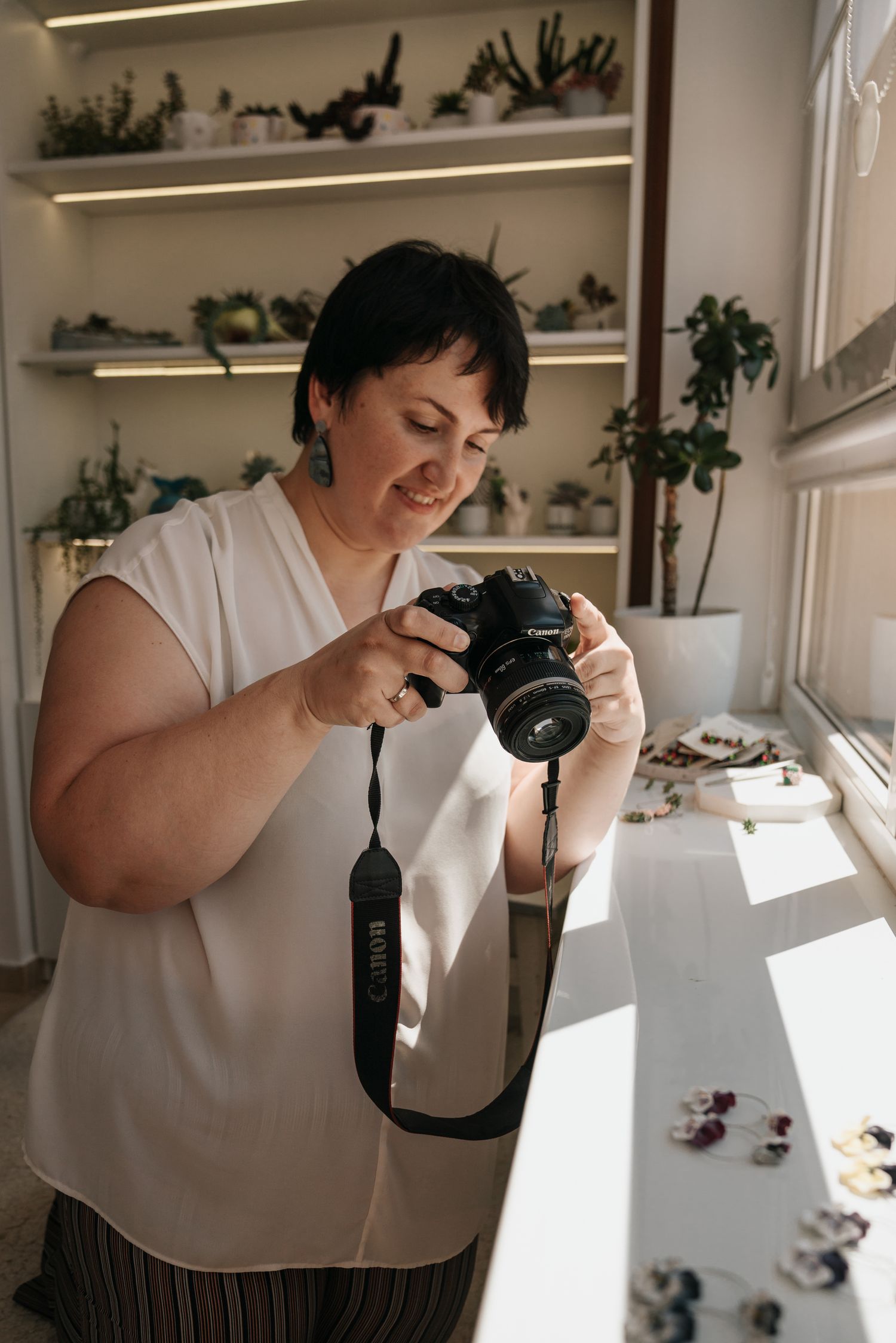 Woman photographer reviewing photos on Canon DSLR camera in bright studio with plant shelves and jewelry display.