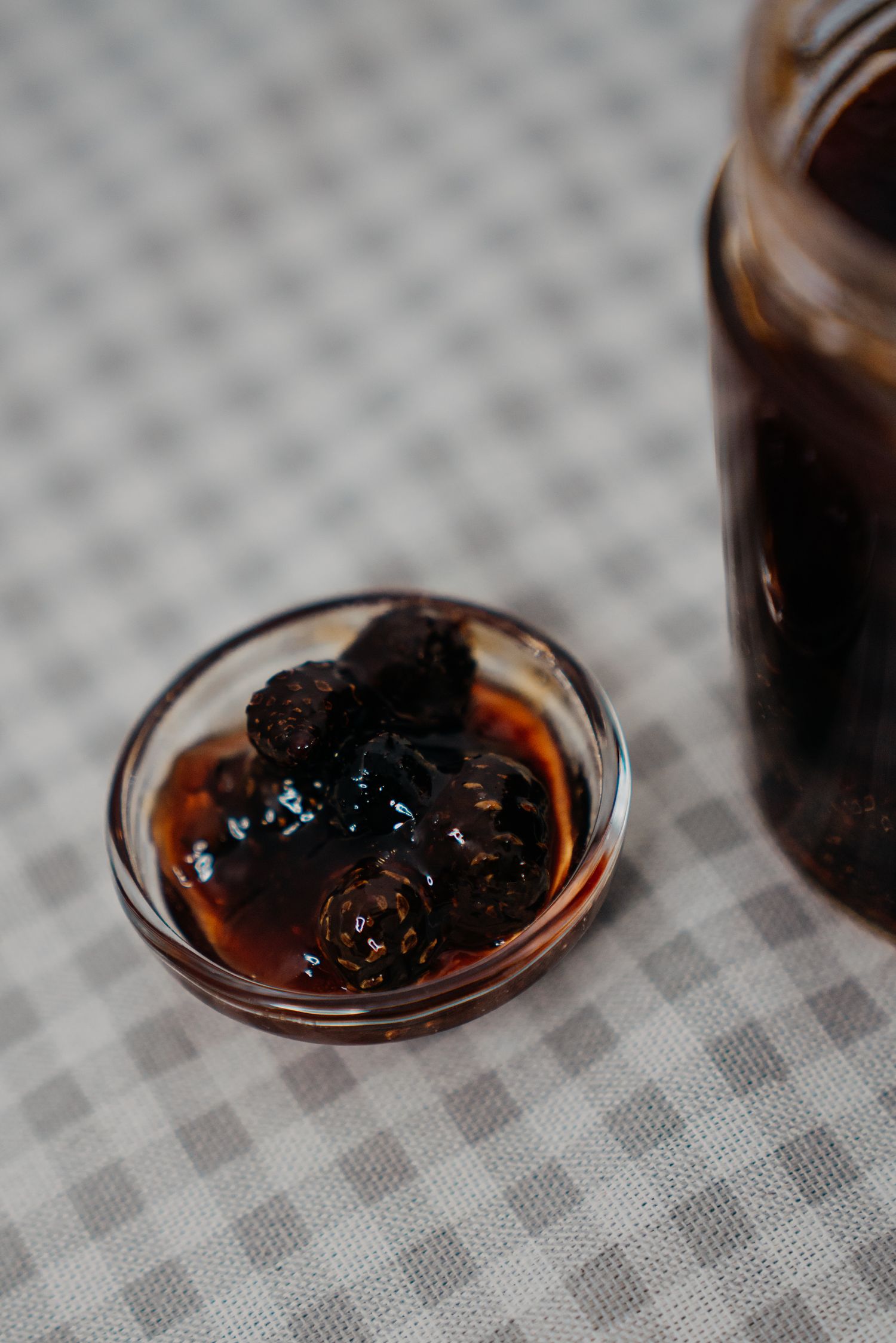 Small glass bowl filled with dark mulberry jam in syrup beside a jar on a checkered tablecloth.