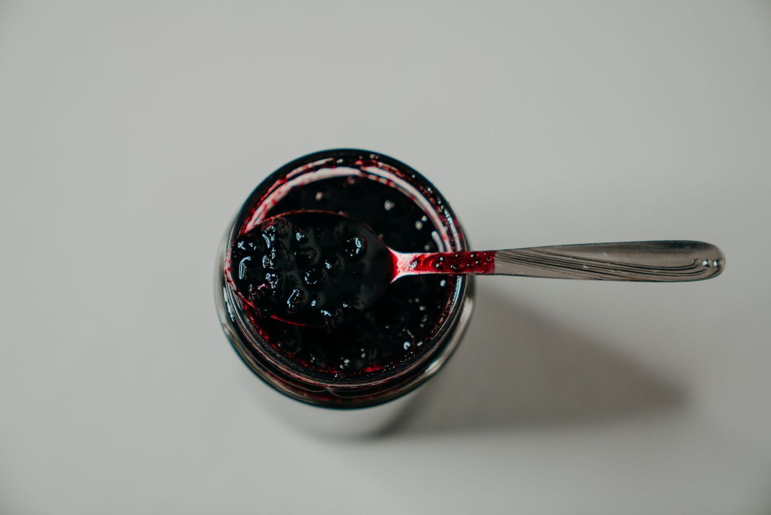 Top view of an open glass jar filled with dark berry jam, with a silver spoon resting inside on a white background.