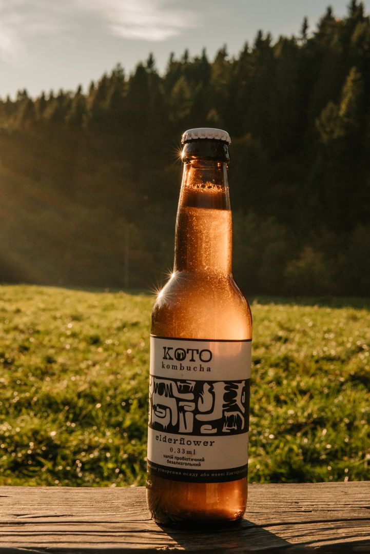 A bottle of Koto Kombucha Elderflower sits on a wooden table with a sunny green meadow and forest backdrop.