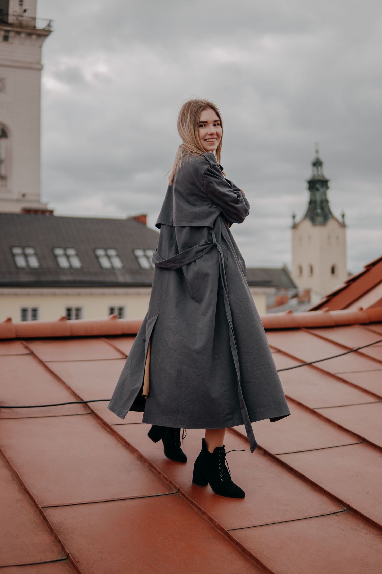 Smiling blonde woman in long gray trench coat posing on red tile rooftop with historic church backdrop.