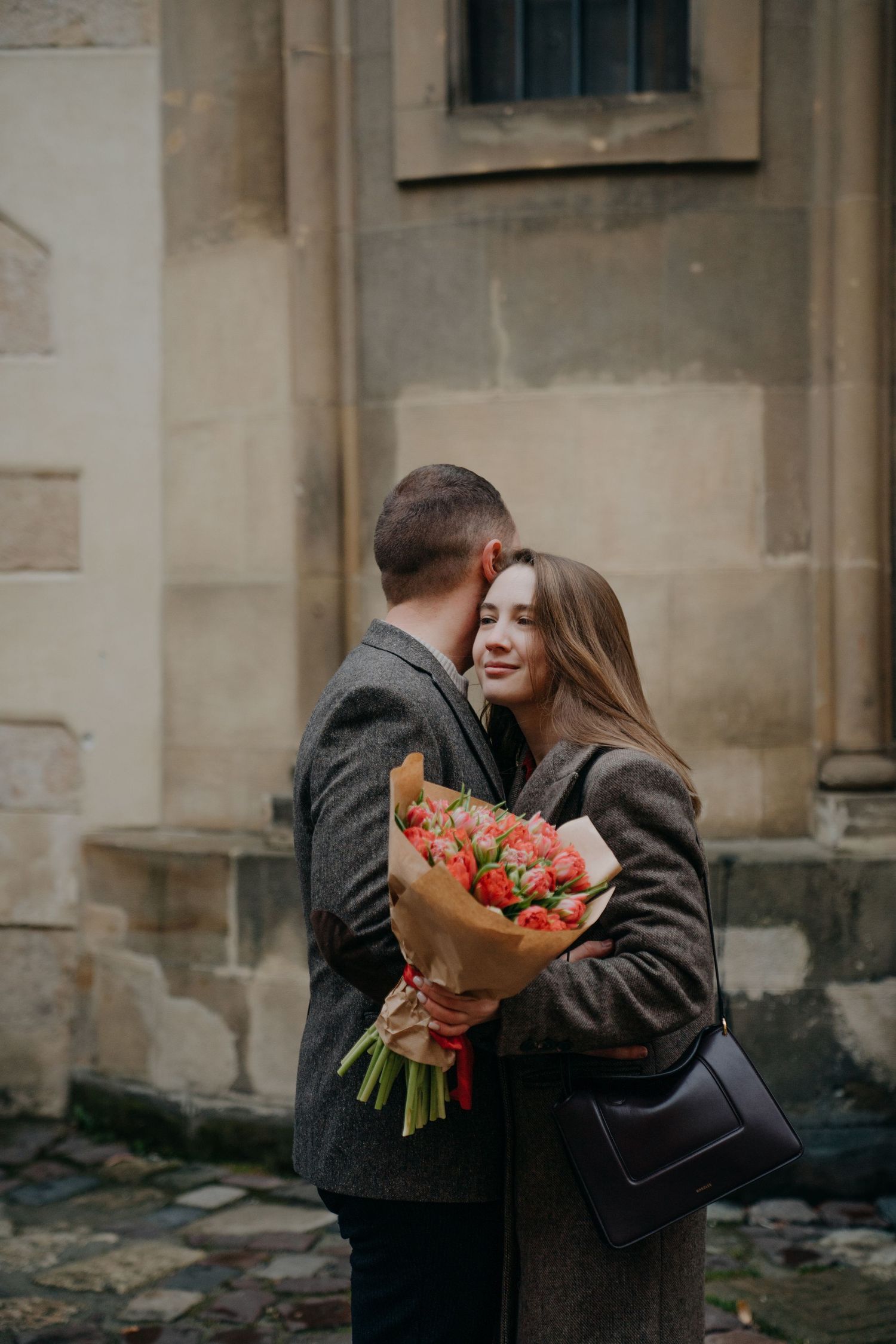 Couple embracing on cobblestone street, woman holding bouquet of red tulips wrapped in kraft paper.