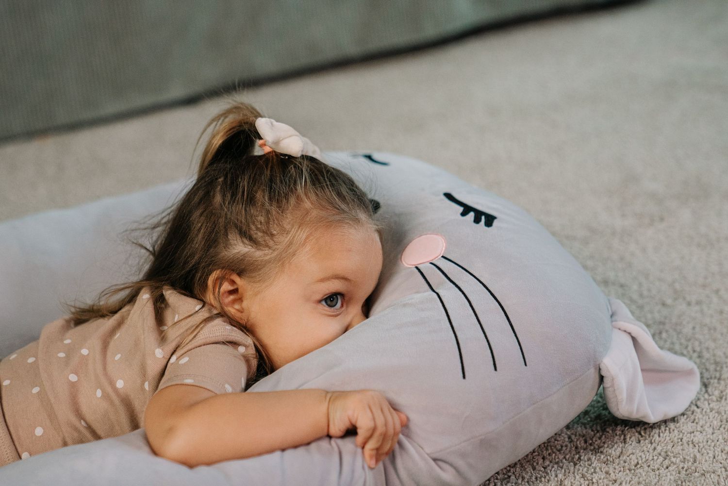 Young girl lying on floor hugging a gray cat-shaped stuffed animal pillow.