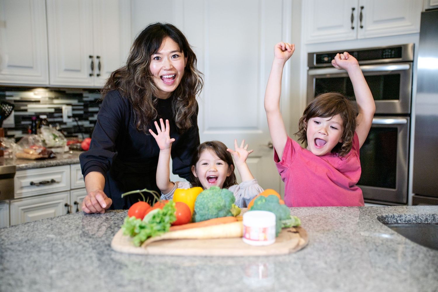 Woman and two excited children posing with fresh vegetables on kitchen counter, celebrating healthy eating together.