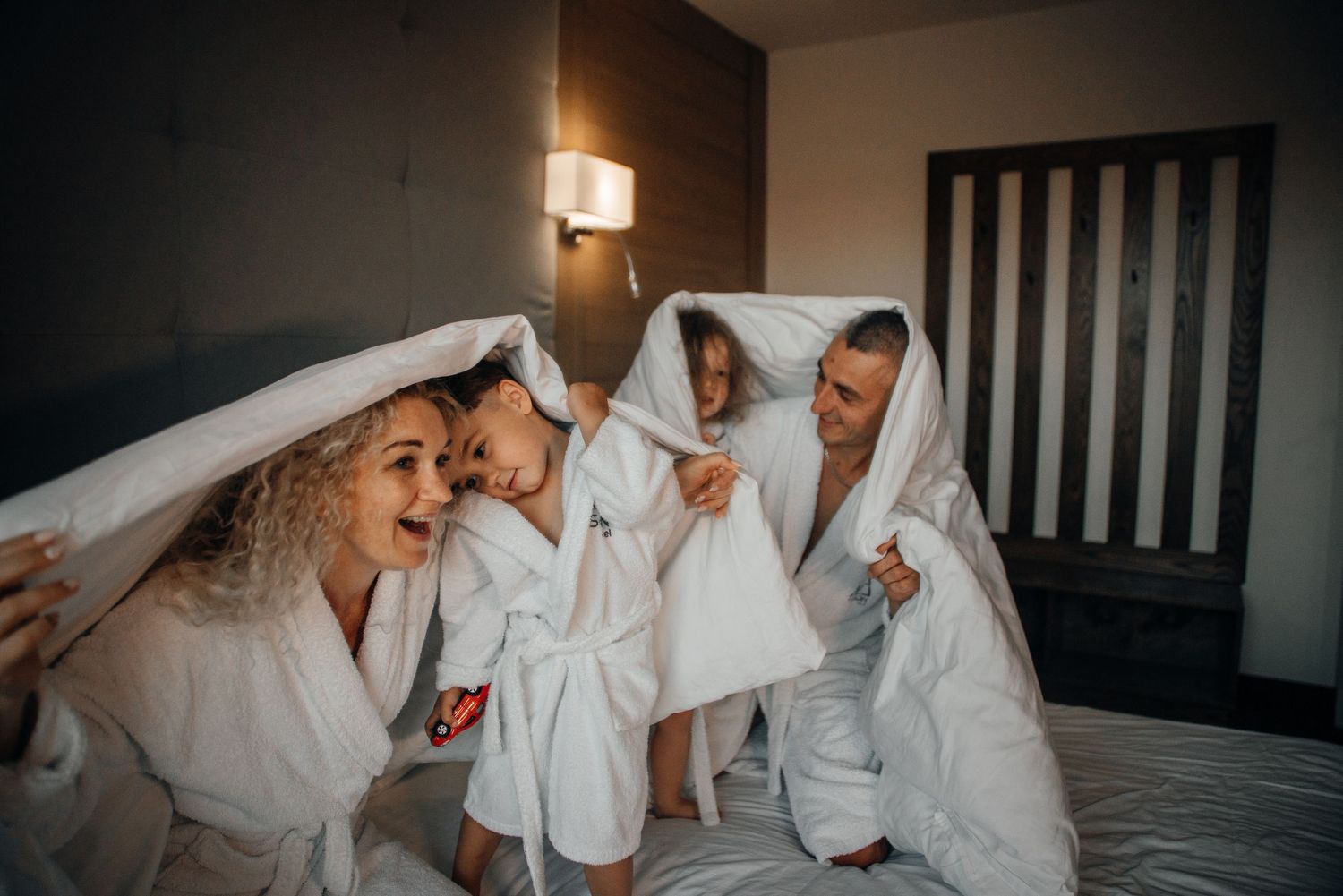 Family in white robes laughing under a blanket on a hotel bed, enjoying a fun and cozy moment together.