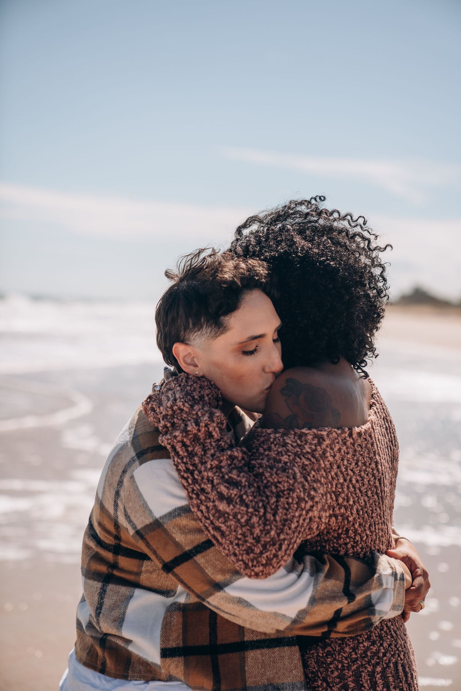 Two people share an intimate embrace on a beach with waves and cloudy skies in the background.