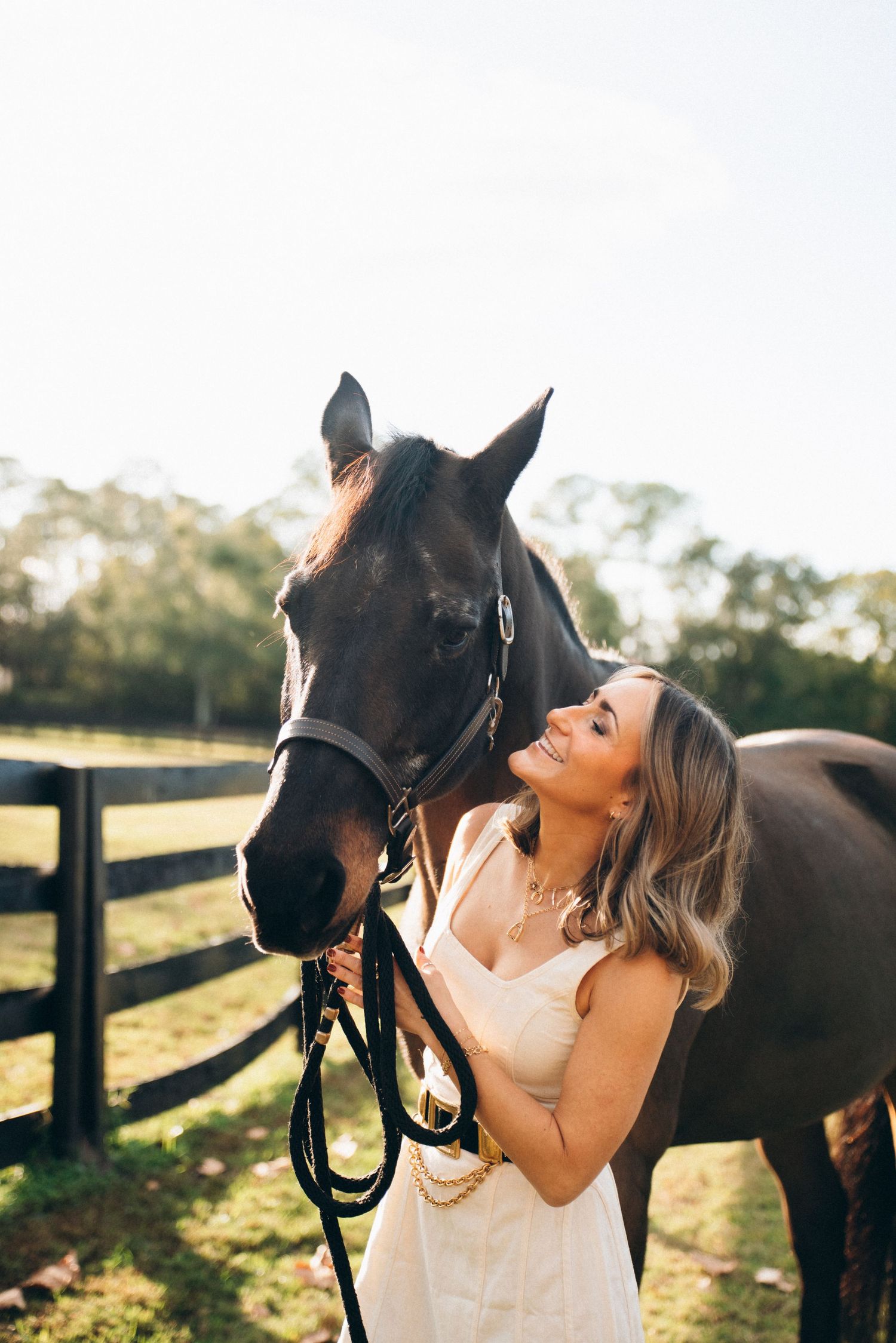 A beautiful dark horse and person in a white dress share a tender moment in a sunlit pasture with wooden fencing.
