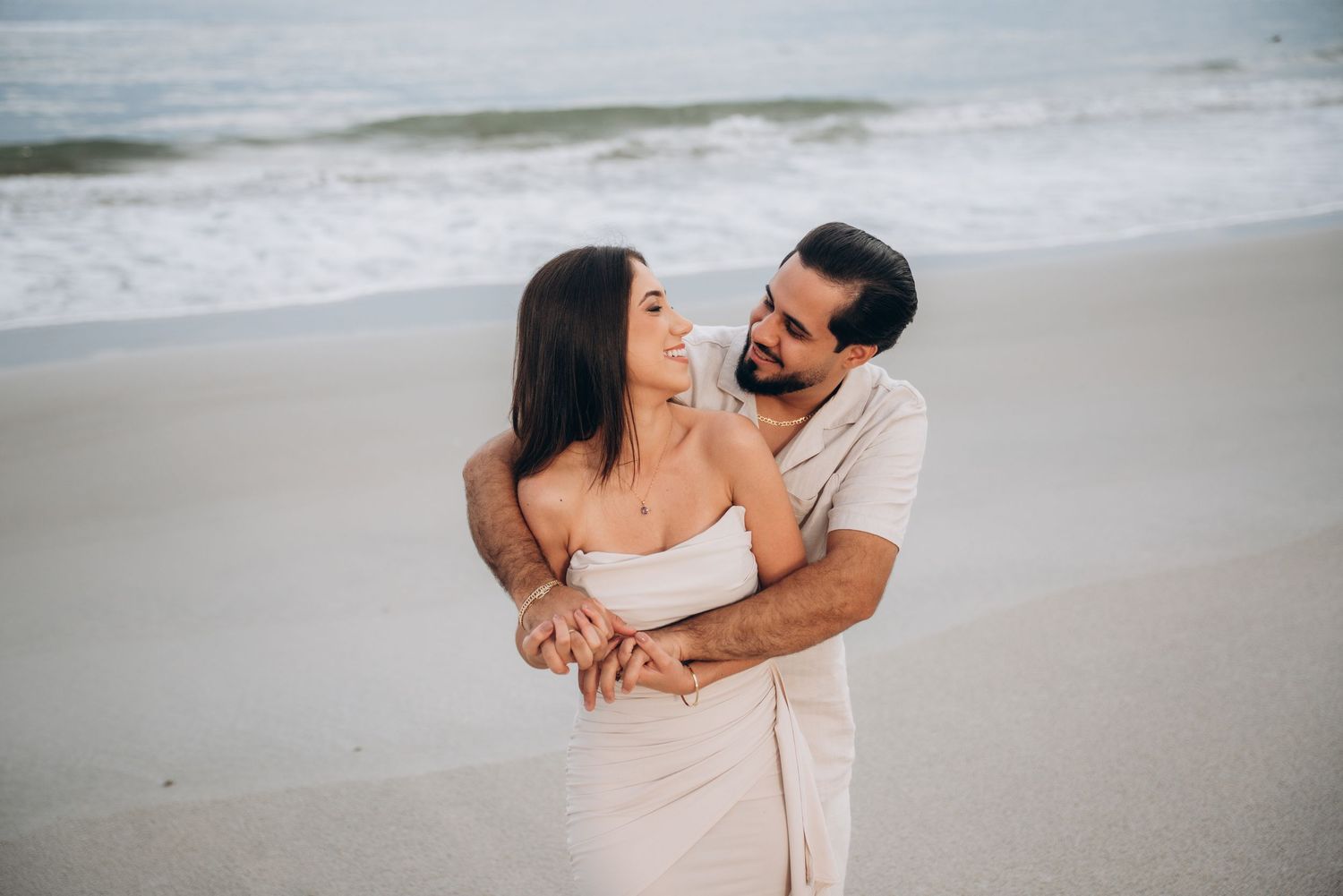 Couple in coordinated beige outfits embracing on empty beach during cloudy day.