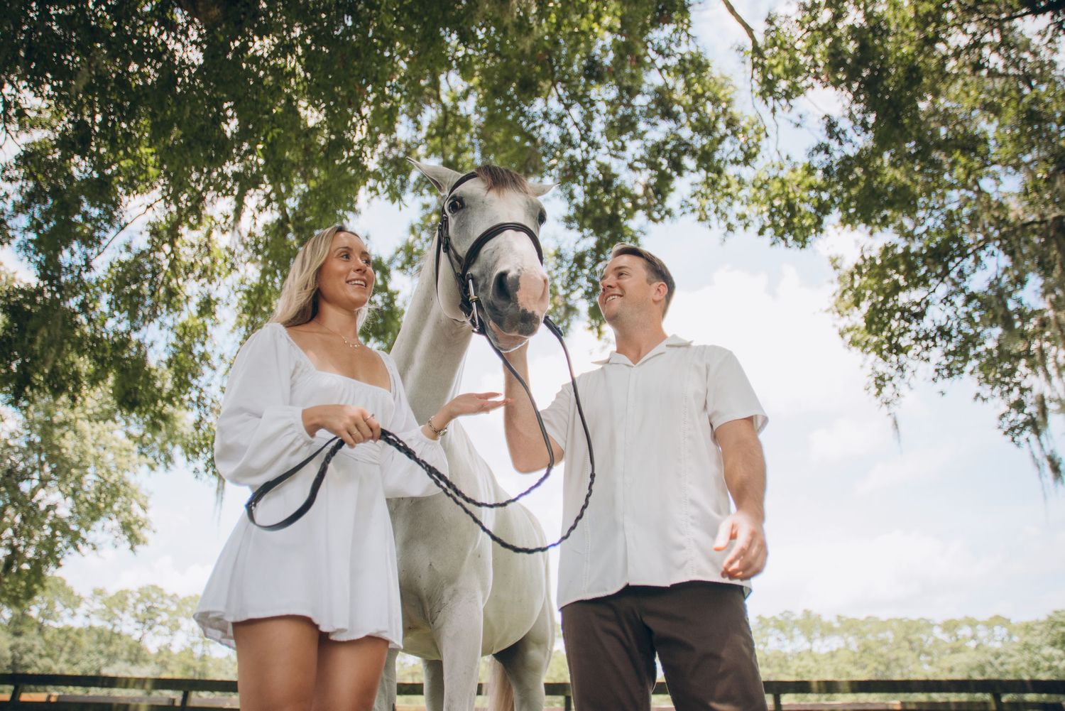 A couple poses with their beautiful white horse in a grassy field with wooden fences.