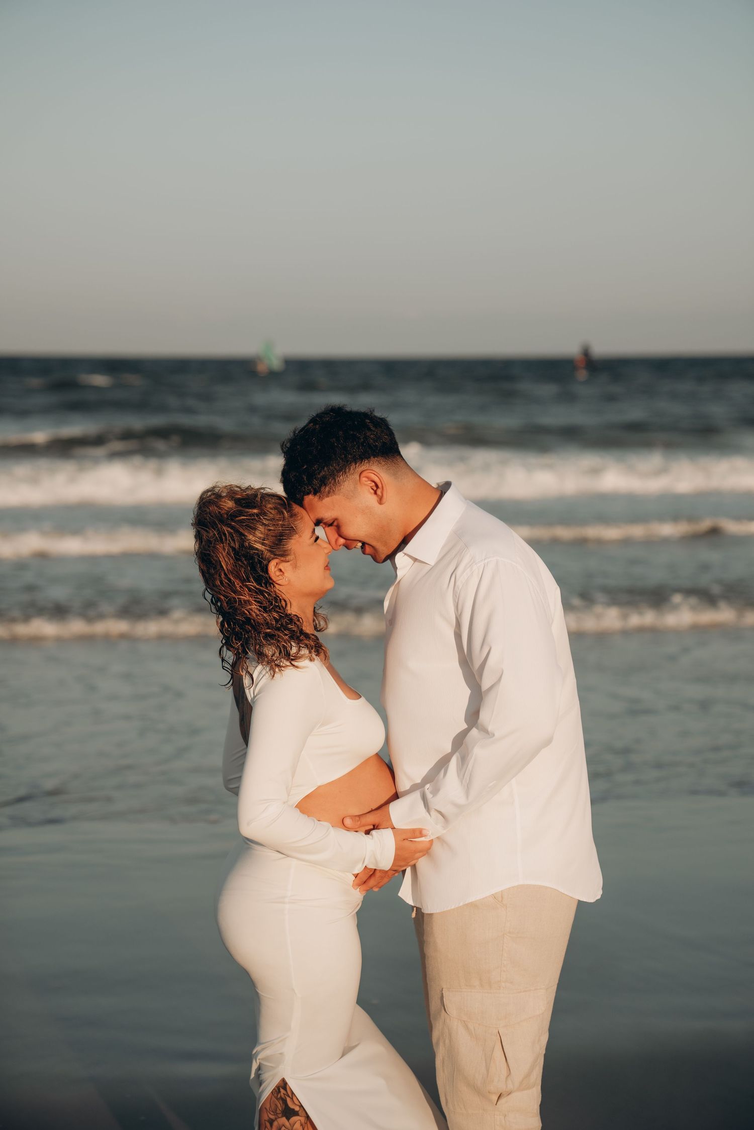 A couple in matching white outfits shares romantic moments during their beach maternity session.