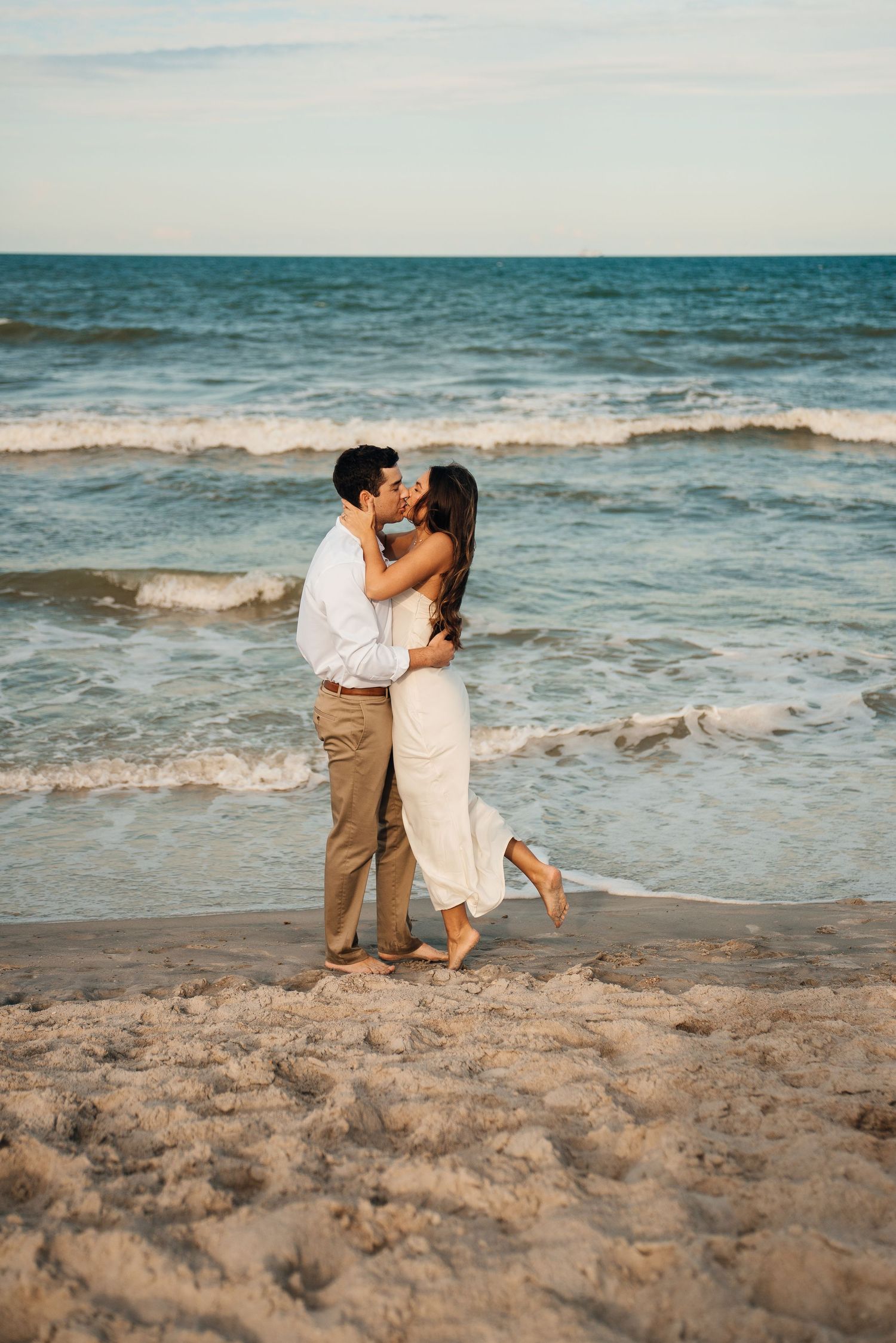 A romantic couple embraces on a beach at sunset with waves crashing behind them.