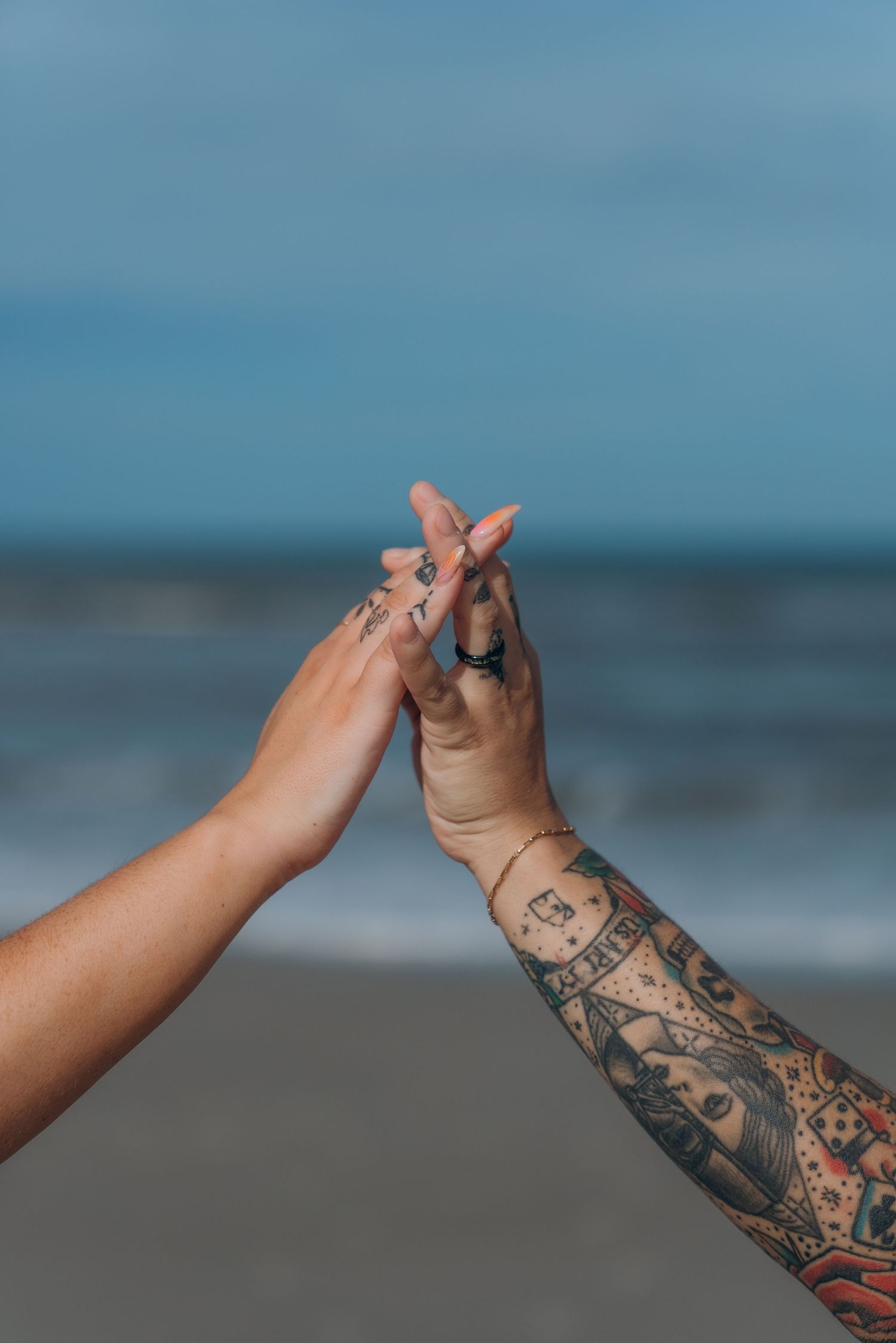 Tattooed arms making heart shapes on a beach with ocean and blue sky in background.