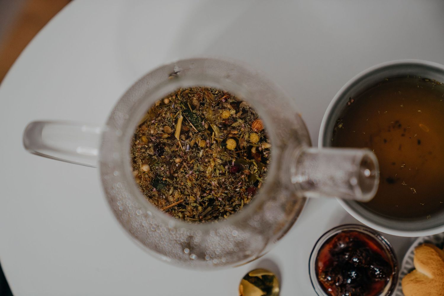 Glass teapot with loose leaf herbal tea blend beside a cup of brewed green tea on a white table.