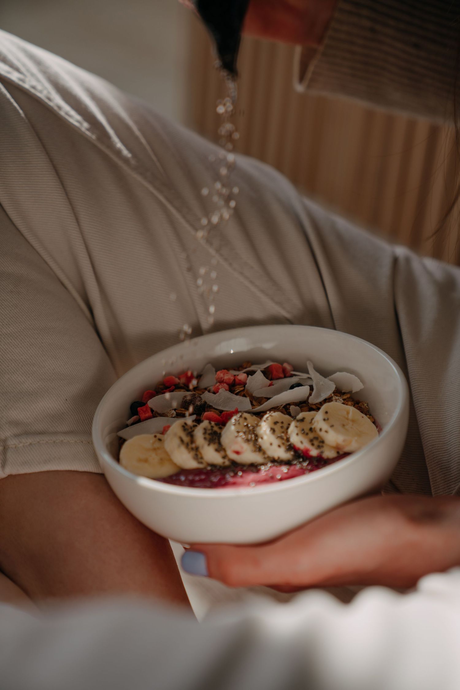 Close-up of person in beige outfit holding a smoothie bowl topped with banana, berries, and seeds.