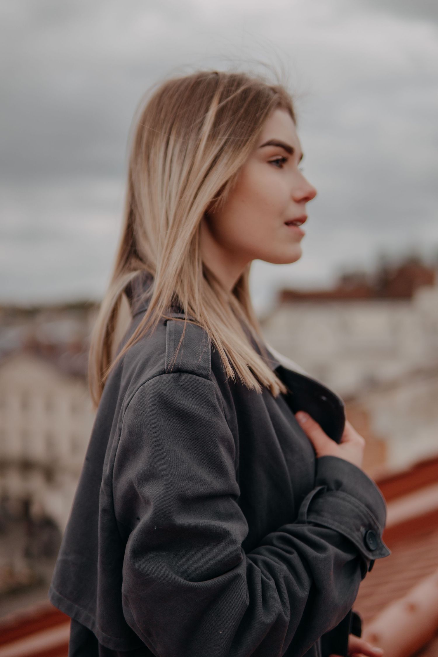 Side profile of blonde woman in dark gray jacket standing on rooftop with cloudy urban skyline behind her.