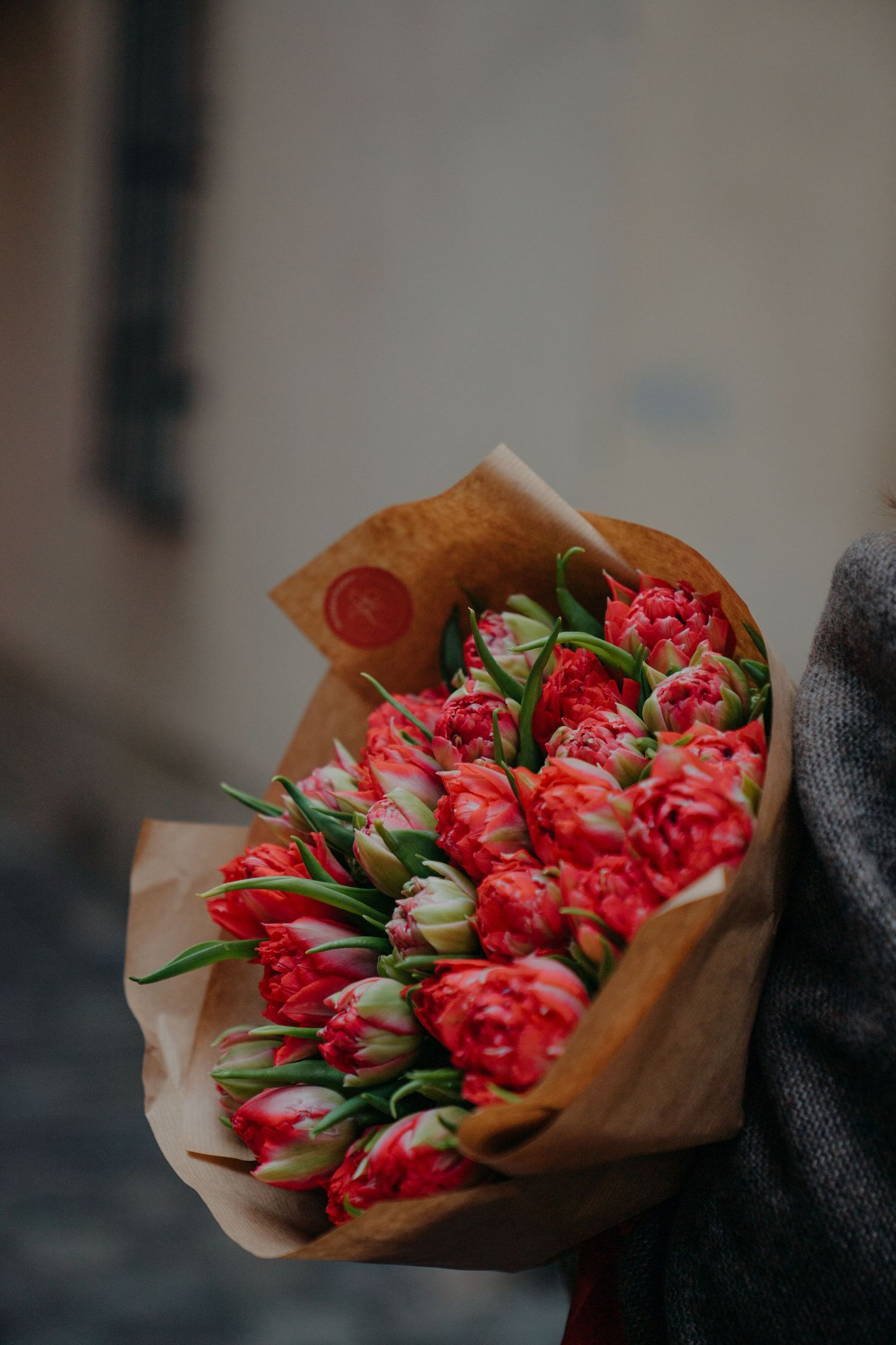 A person holds a large bouquet of vibrant red and pink tulips wrapped in brown kraft paper outdoors.