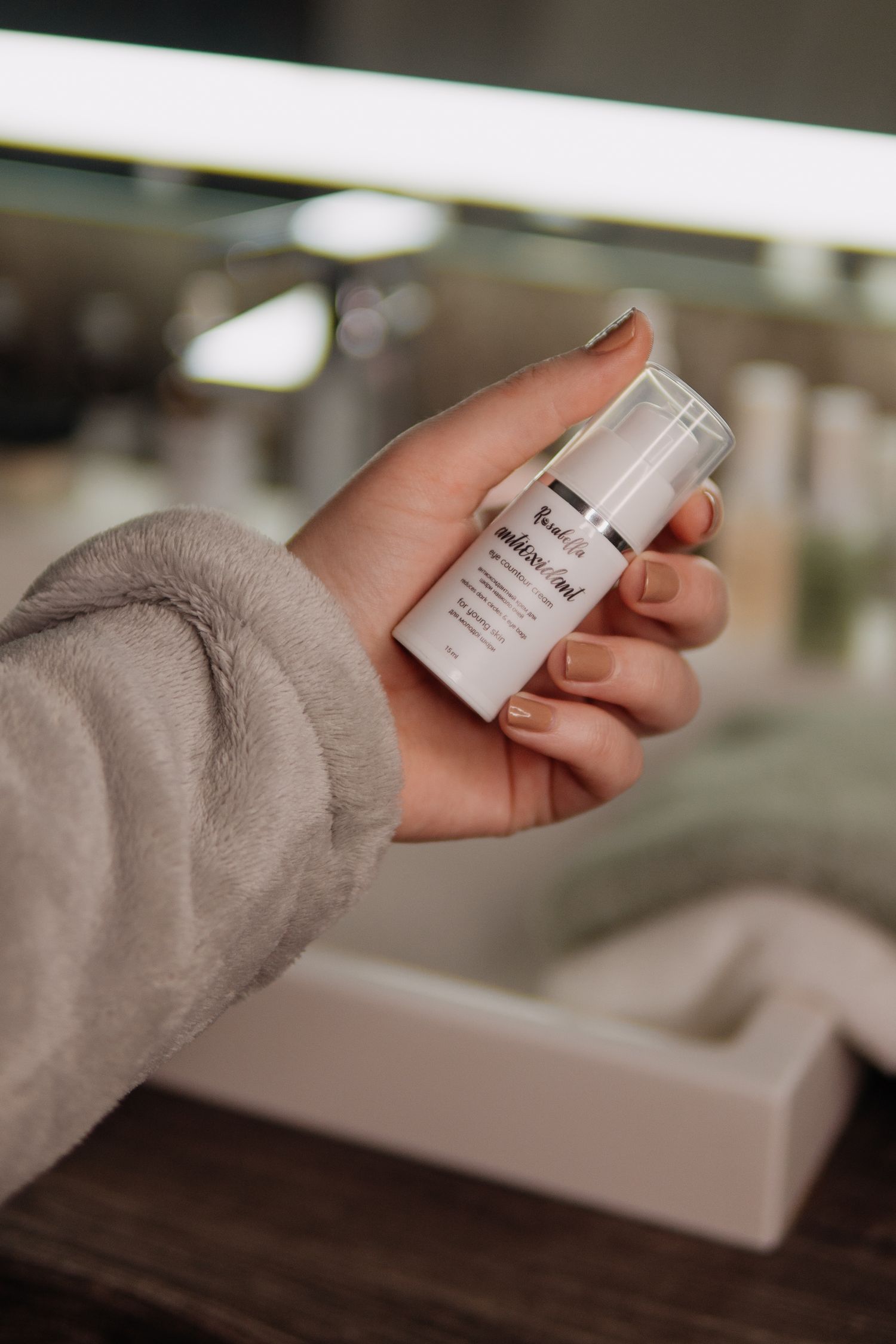 Woman in gray robe holding a white skincare serum bottle in a modern bathroom setting.