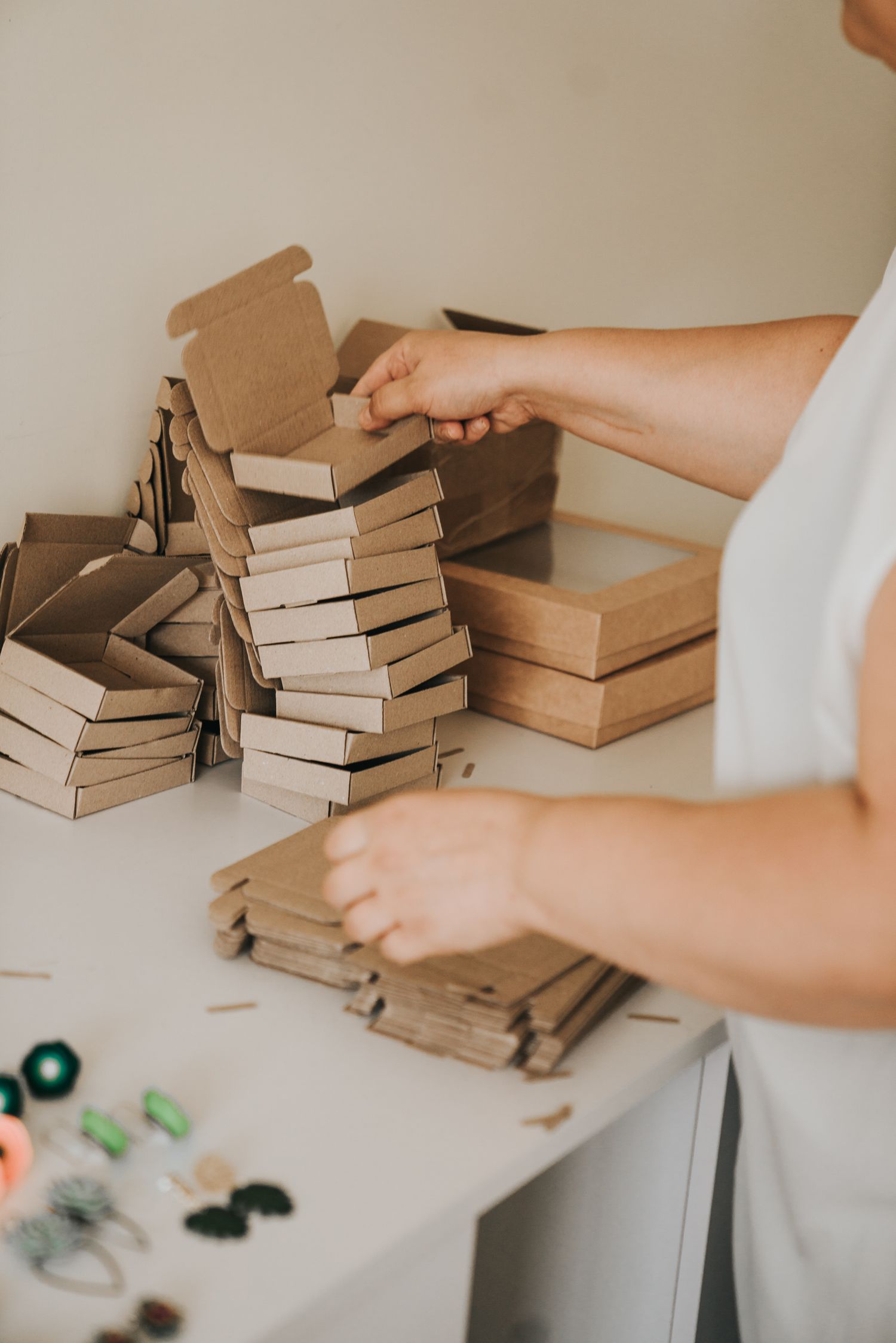 Person assembling stacks of small brown kraft cardboard boxes on a white table for product packaging.
