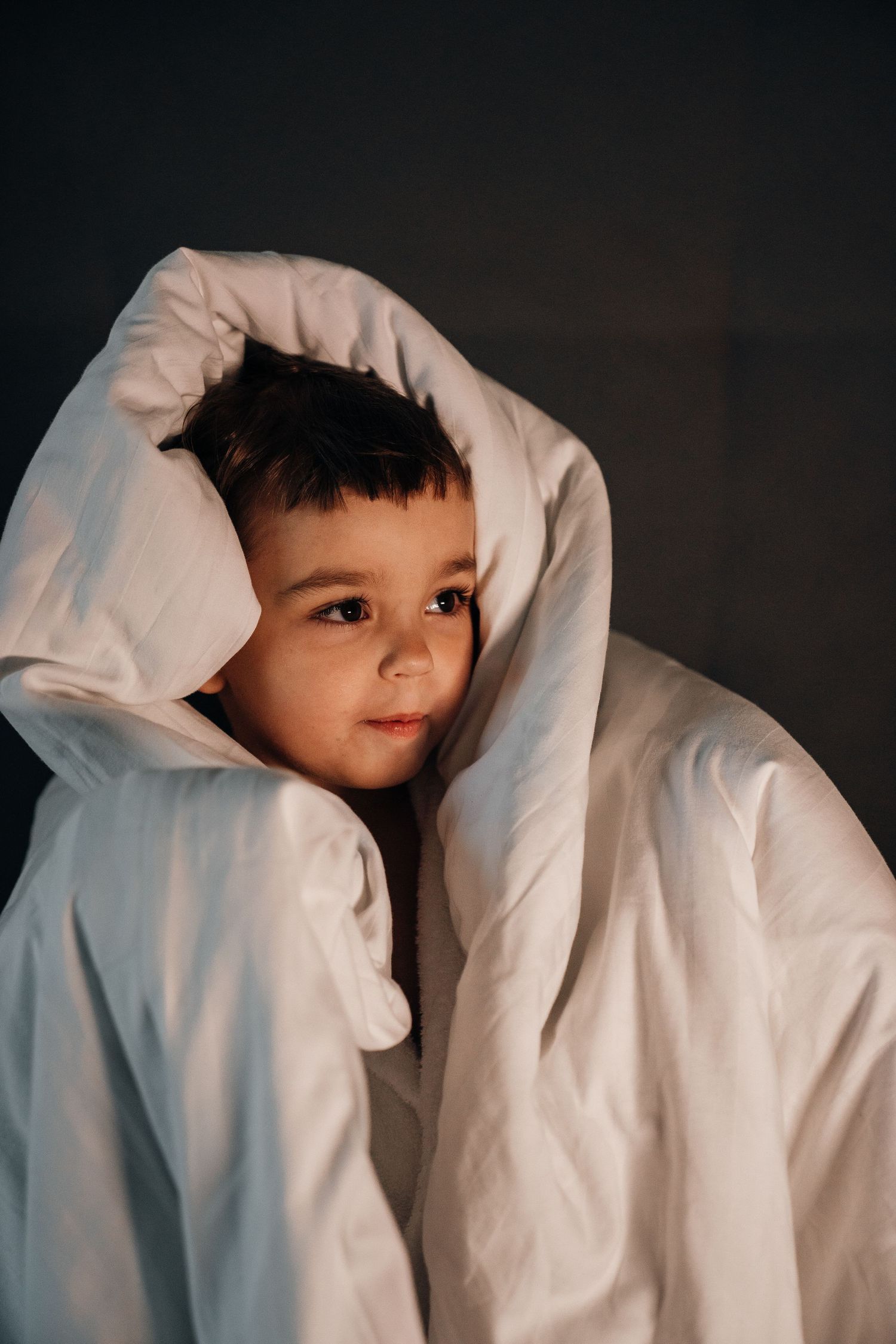 Young child wrapped in a white duvet blanket looking up in warm golden light.