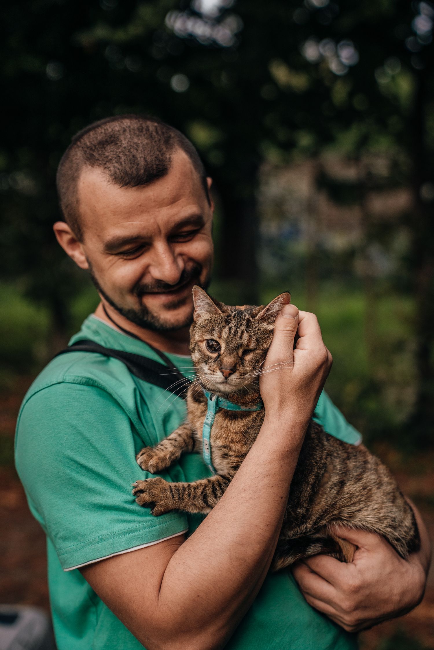 Smiling man in green scrubs gently pets a tabby cat with blue collar outdoors.