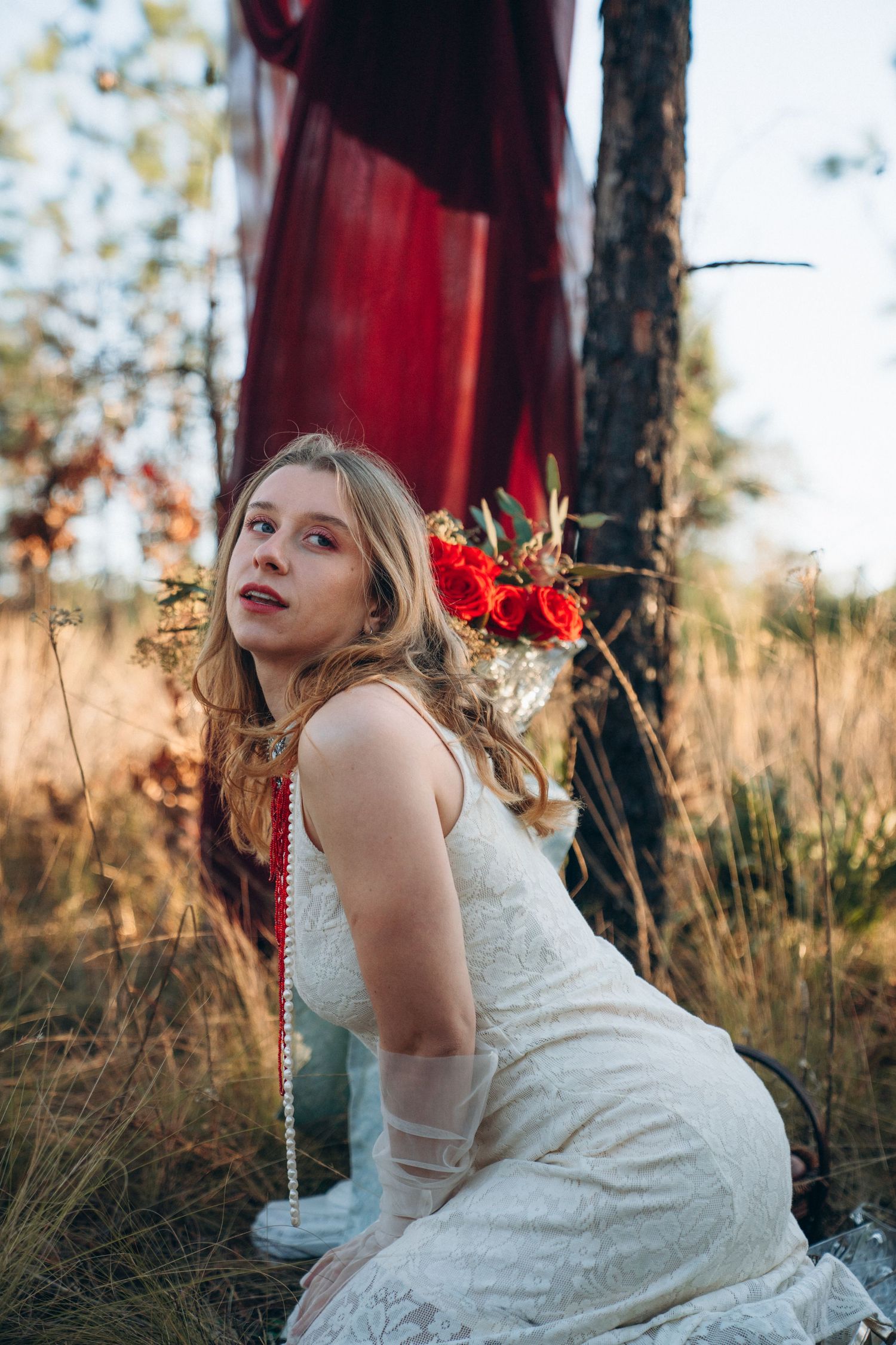 Blonde woman in white lace dress with red flower crown kneeling in golden grass near red draped tree.
