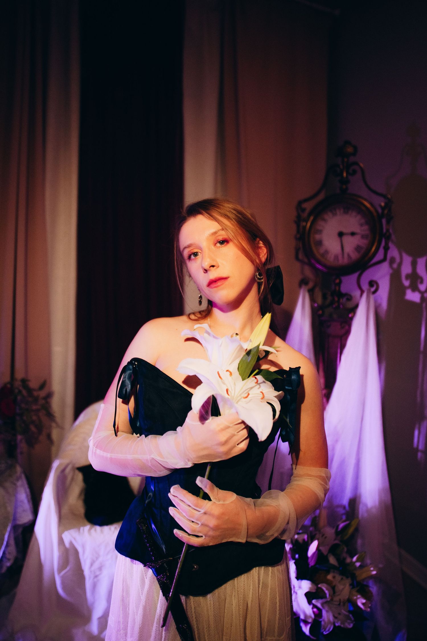 Young woman in black corset holding white lily flowers, wearing white gloves in a dimly lit vintage room.