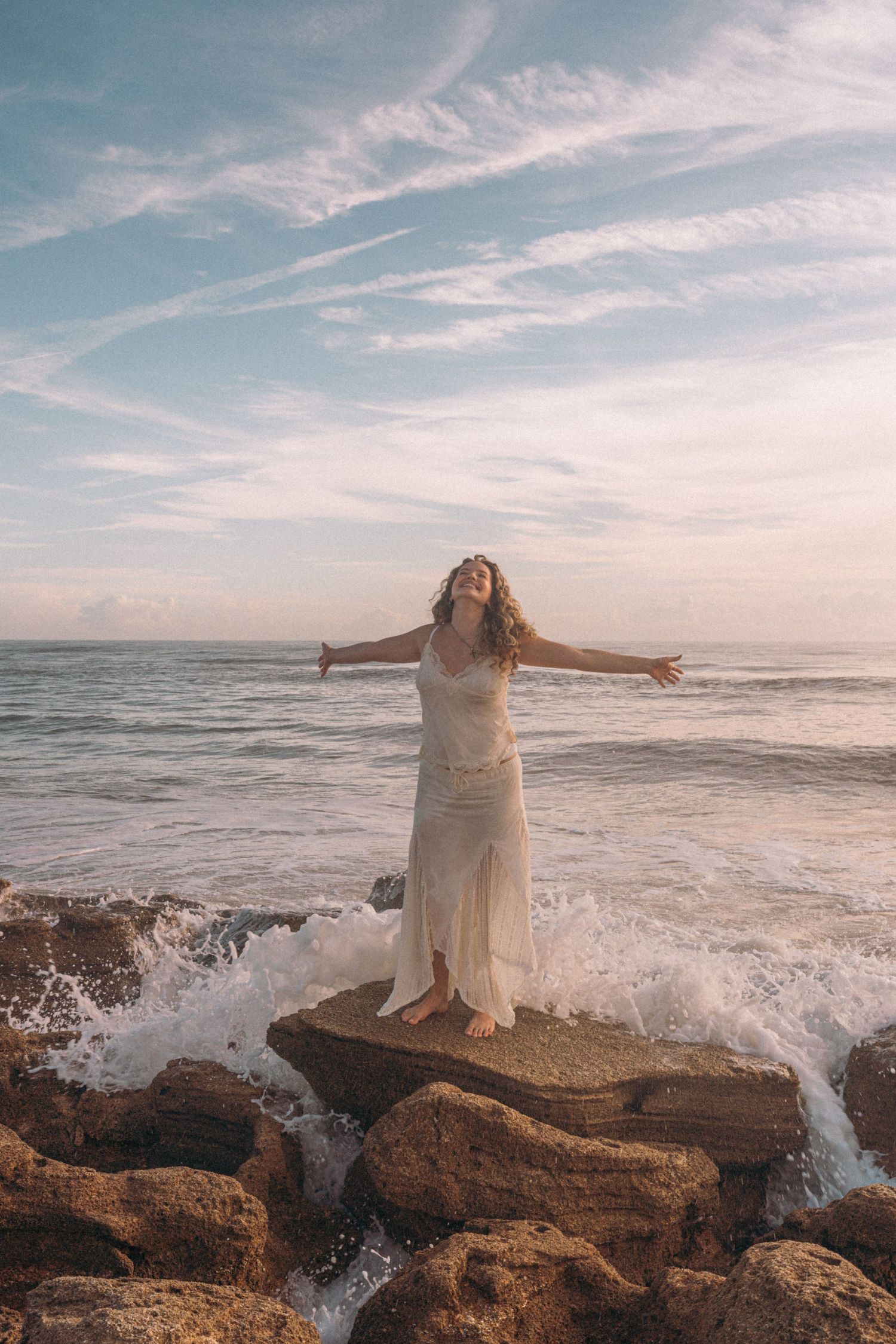 Woman in white dress stands on rocks with arms outstretched as ocean waves crash around her at sunset.