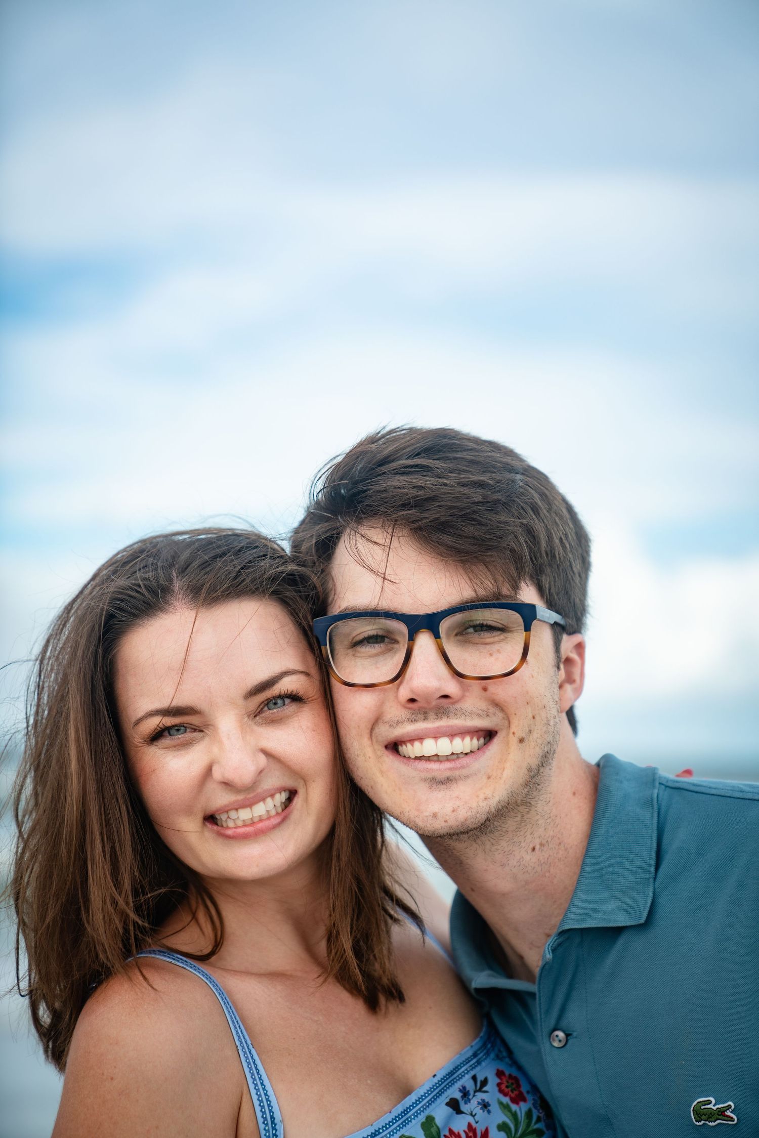 A couple poses for close-up selfies together against a bright blue sky background.