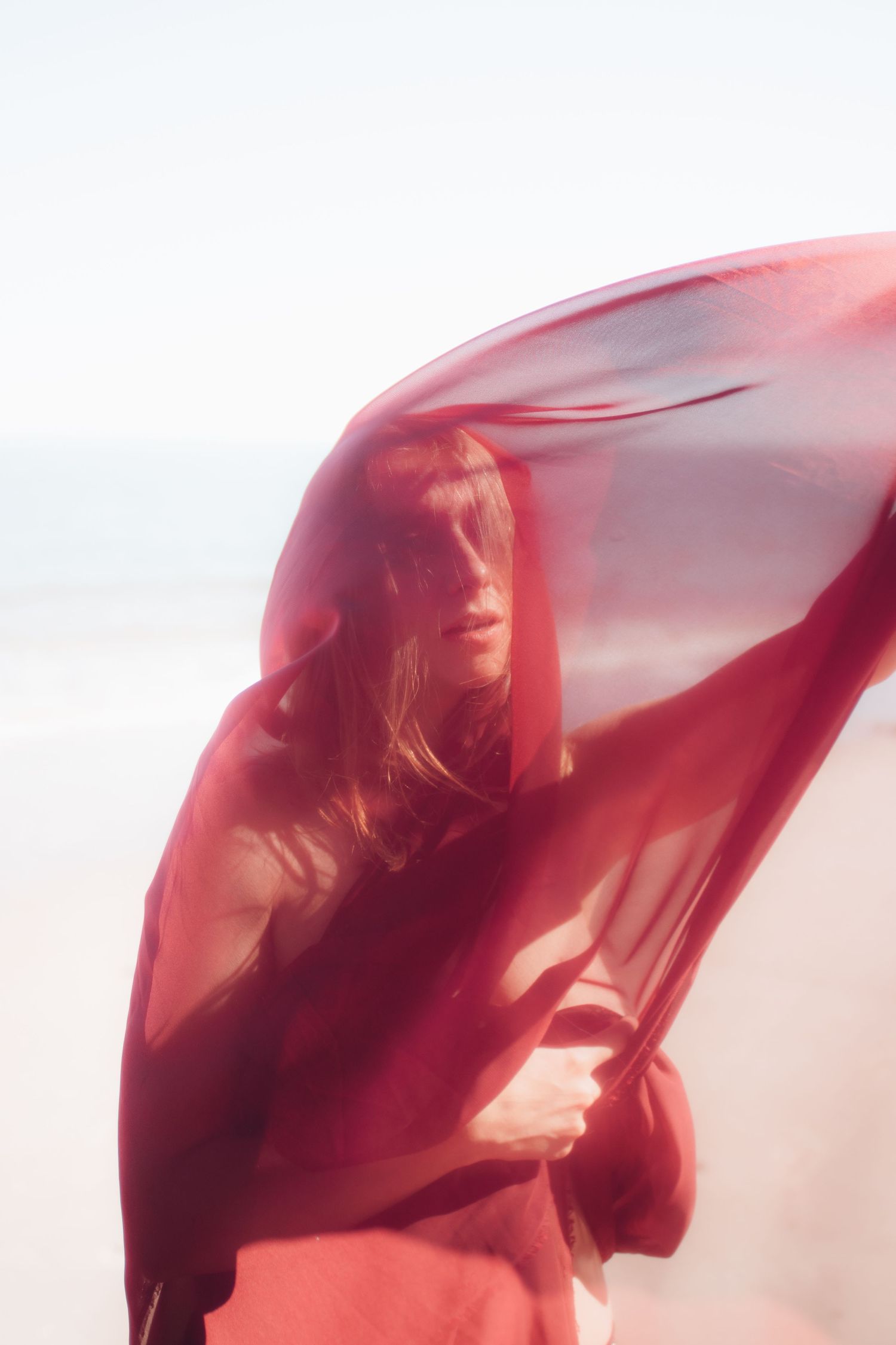 A figure draped in flowing red fabric stands against a bright background creating an ethereal silhouette effect.