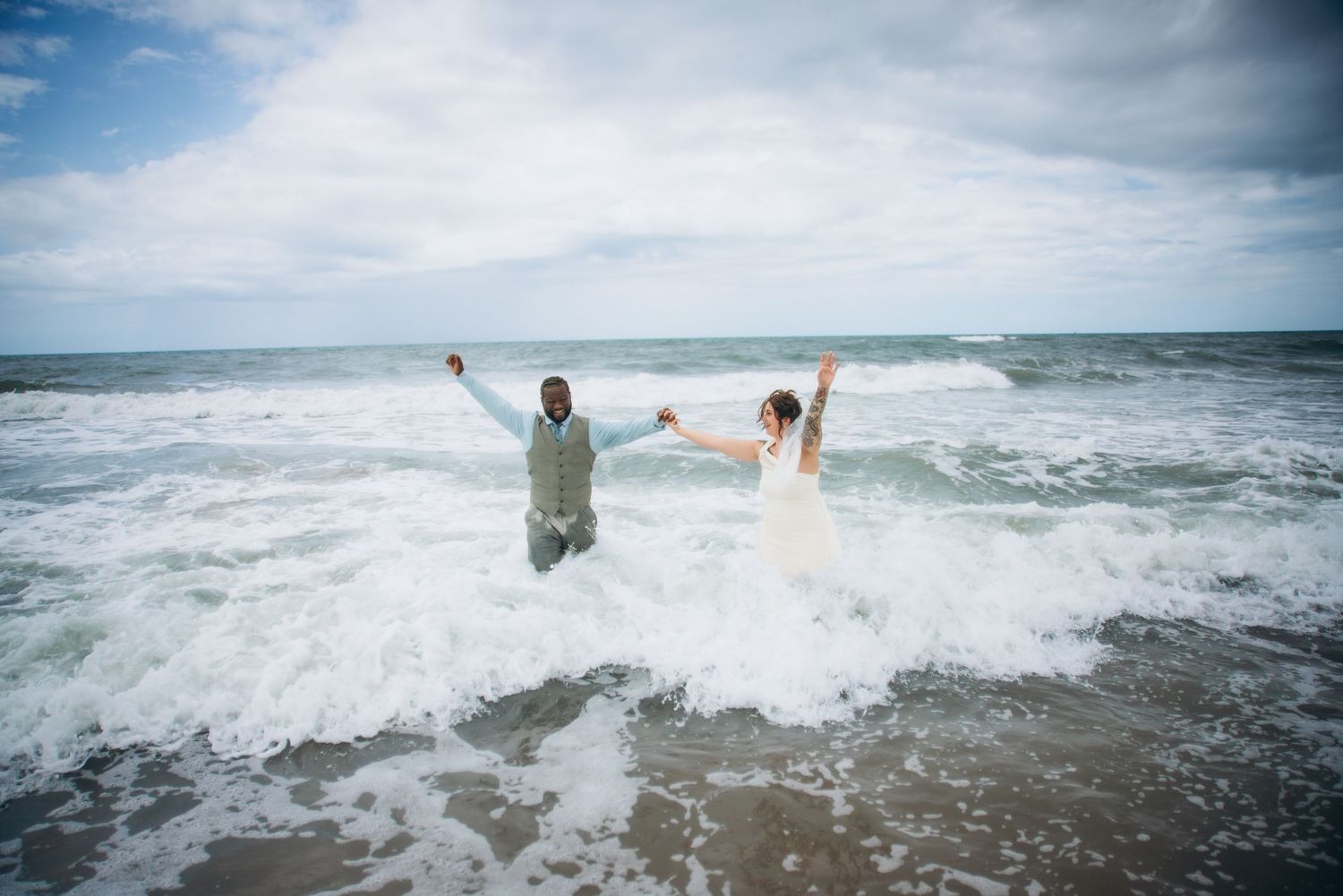 Newlywed couple in wedding attire celebrating in ocean waves on a sandy beach on a cloudy day.