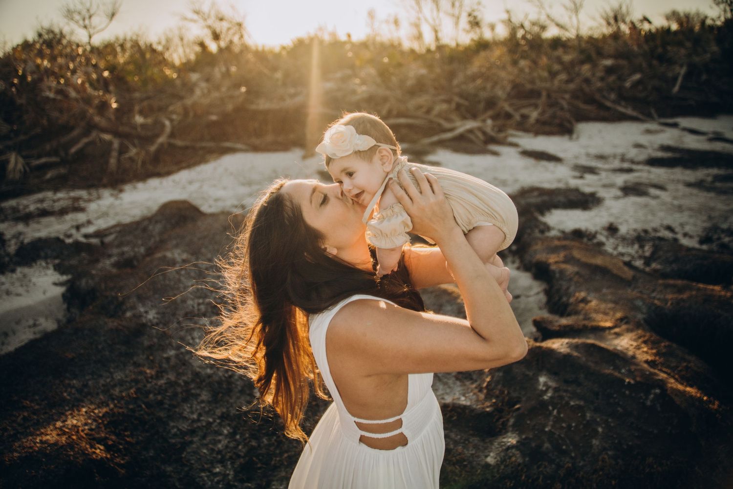Woman in white dress holding a baby up in golden sunset light on rocky terrain.