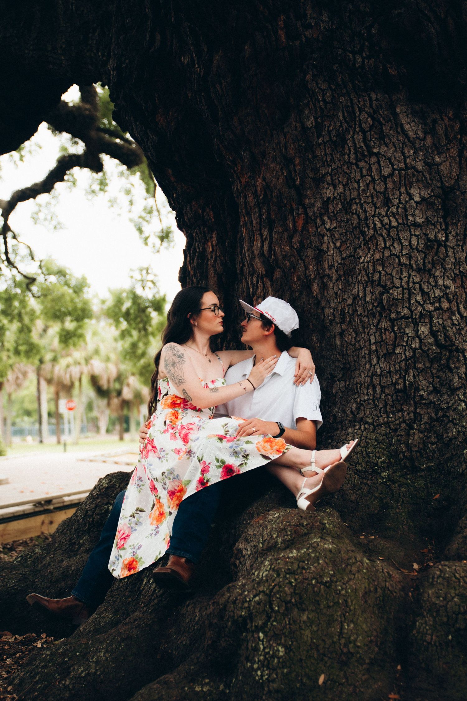 Romantic couple sitting together against massive tree trunk, woman in floral dress, man in white outfit and cap.