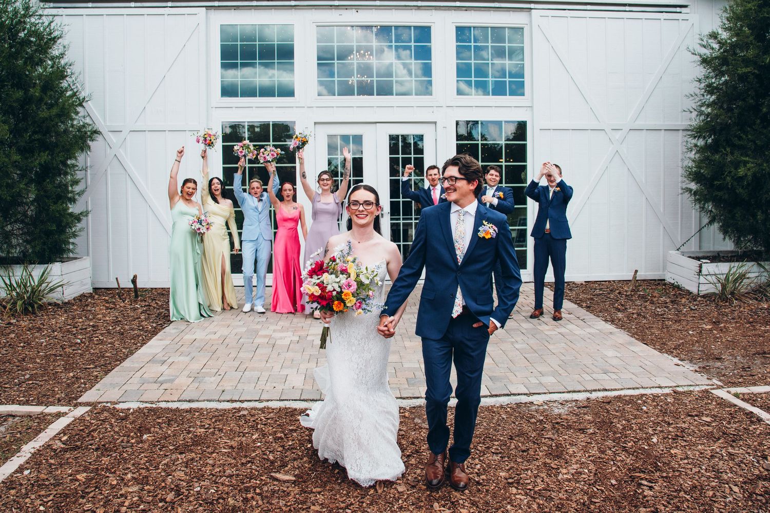 Bride and groom walk hand-in-hand as wedding party cheers behind them outside a white barn venue.