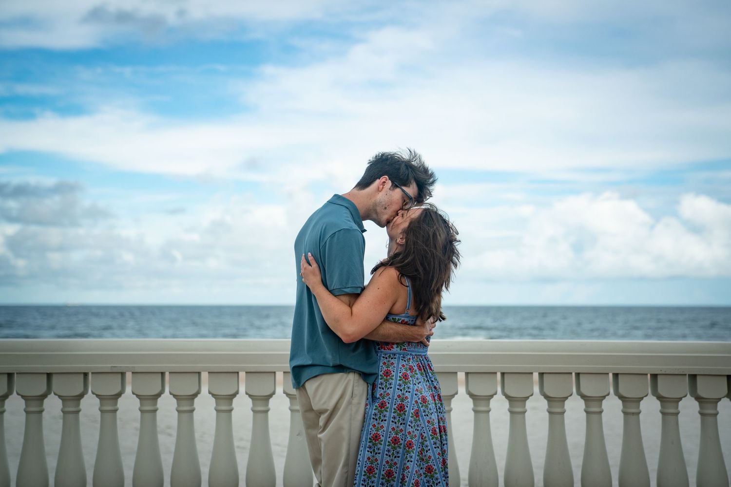A romantic couple shares an intimate moment along a seaside balustrade with ocean and cloudy sky in the background.