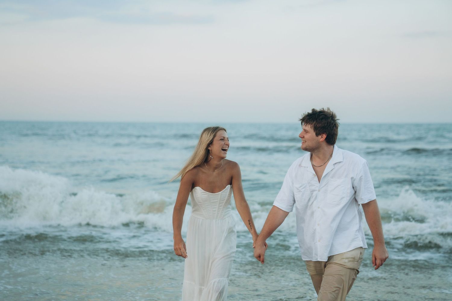 Couple holding hands while walking along empty beach at sunset.
