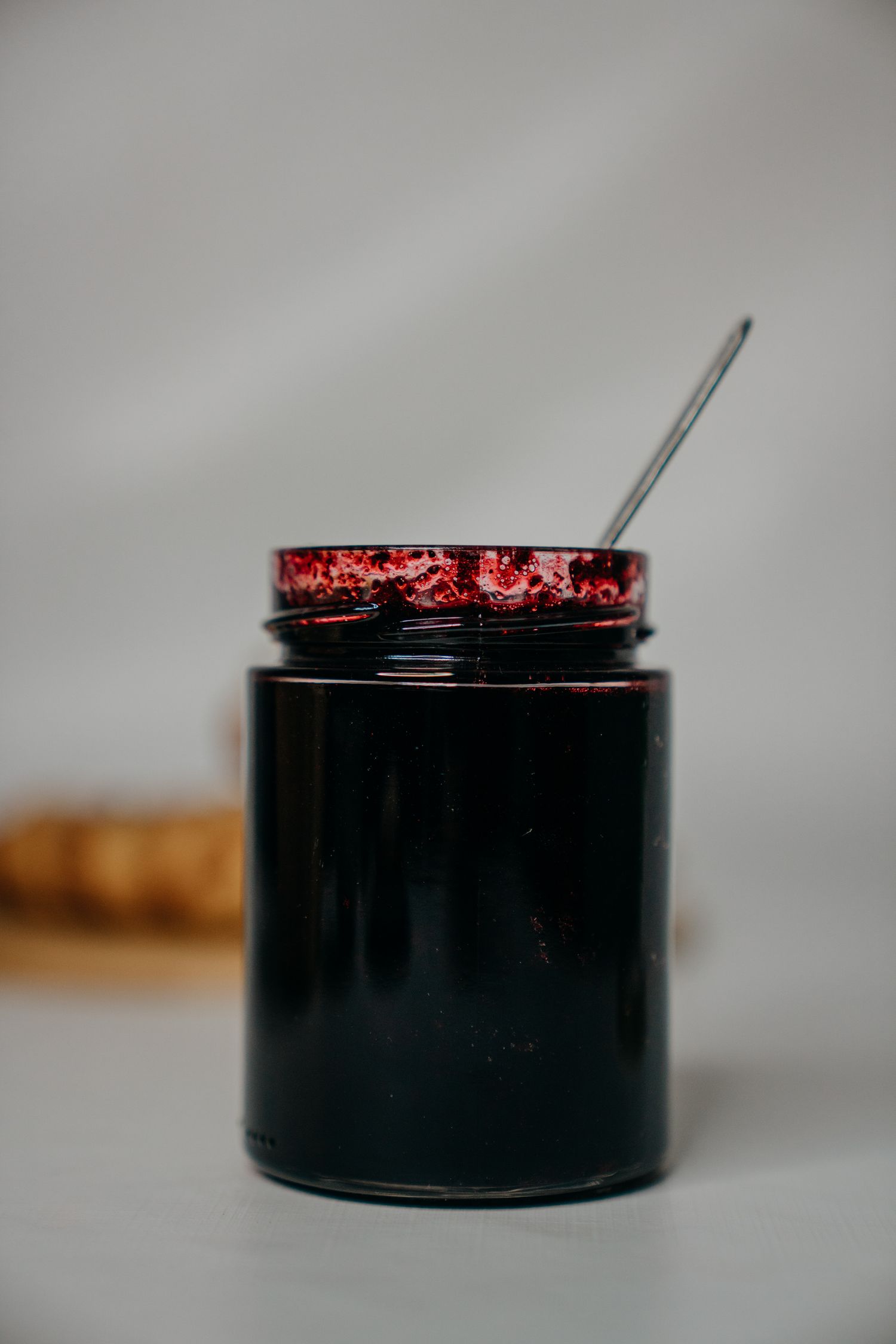 Open glass jar of dark berry jam with a metal spoon on a light gray surface.
