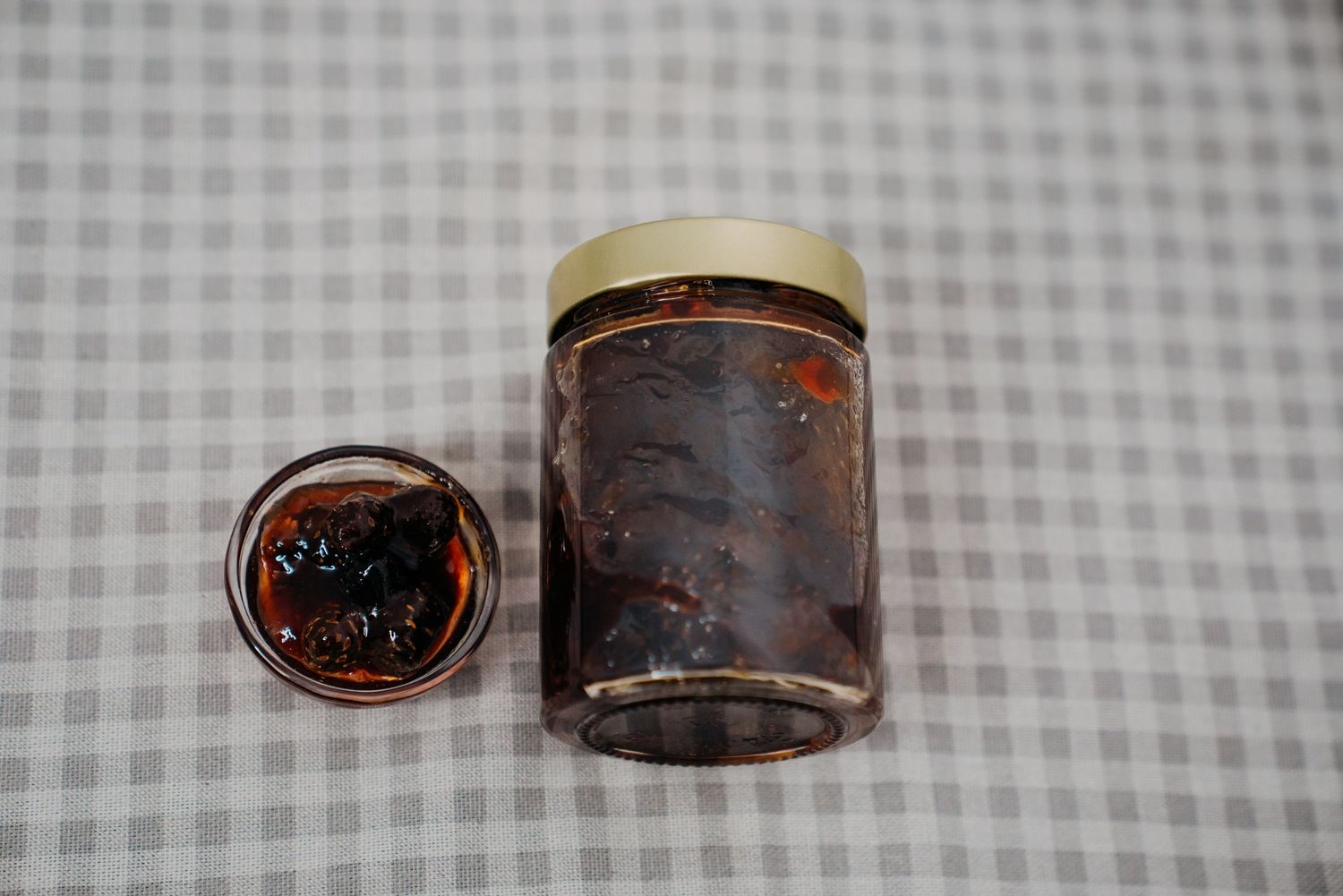 Glass jar of dark fruit jam with lid beside a small open jar on a checkered tablecloth.