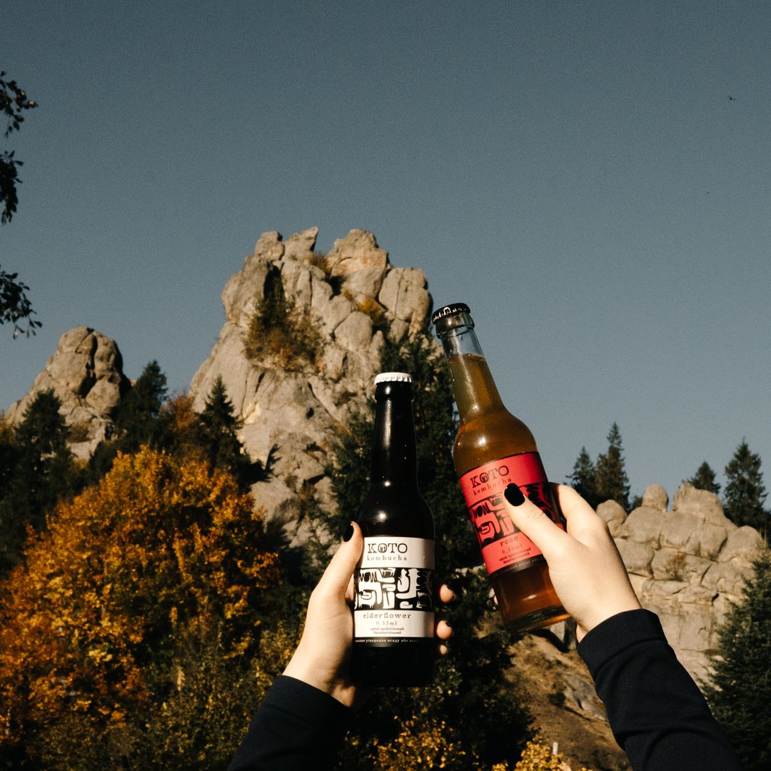 Two hands holding craft beer bottles outdoors with rocky mountain peaks and autumn trees in background.