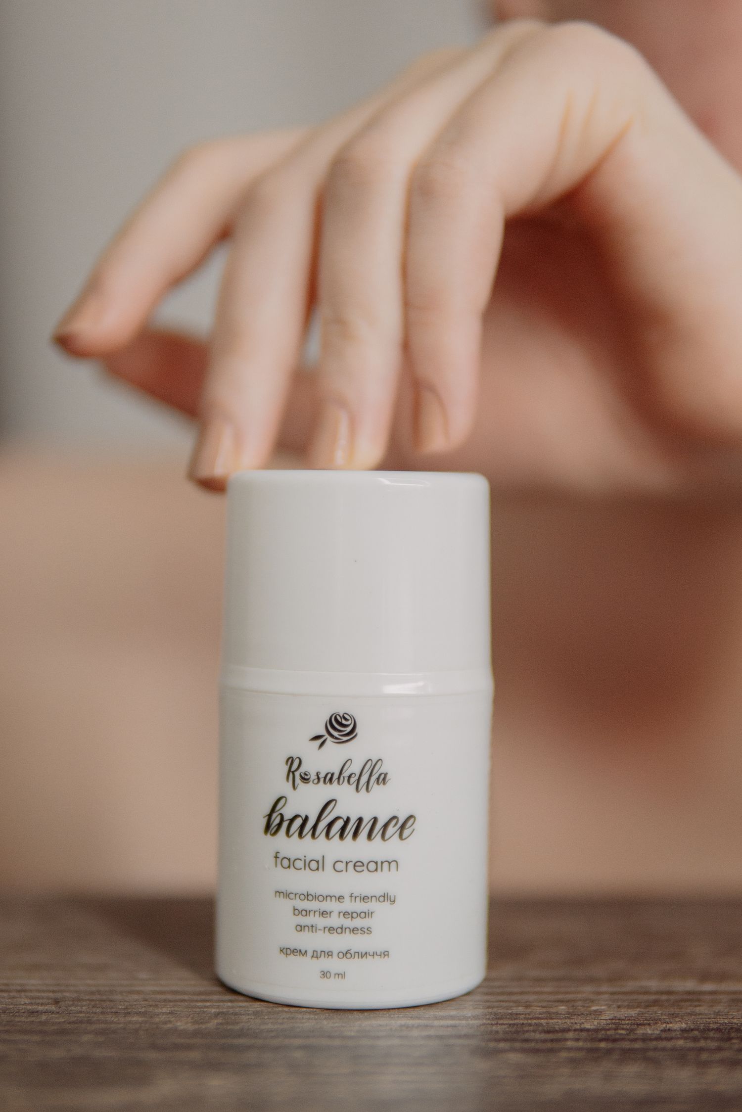 Rusaloffa Balance facial cream in white airless pump bottle on wooden surface with womans hand reaching overhead.
