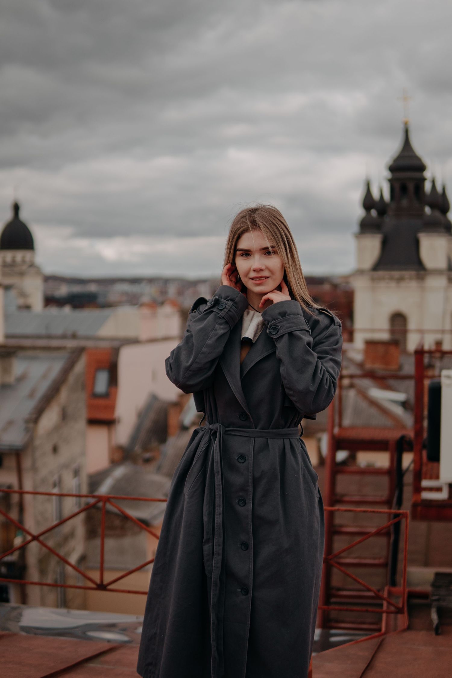 Young woman in dark trench coat poses on rooftop with cloudy city skyline backdrop.