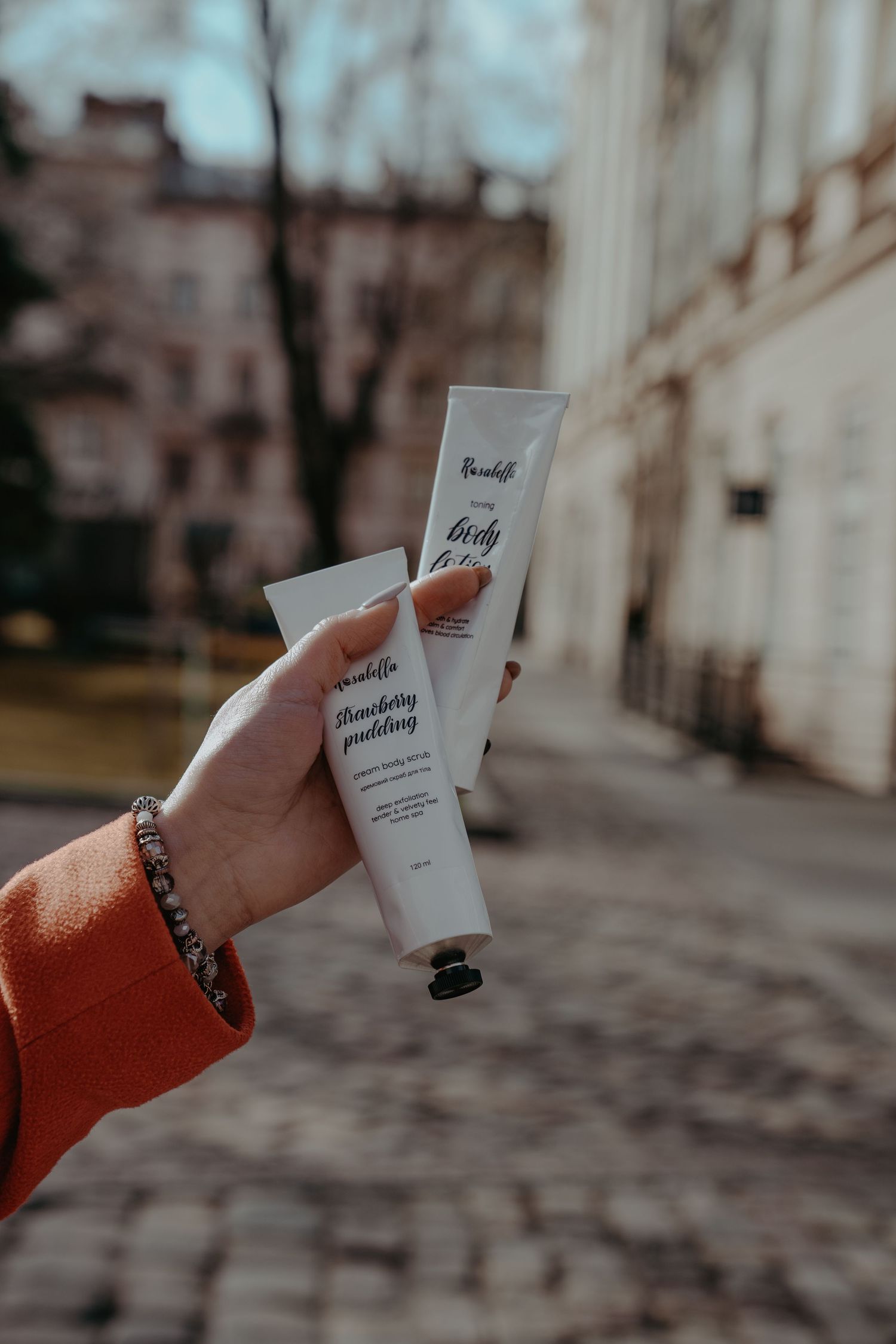Hand holding two Rosabella body cream tubes outdoors on a city street.