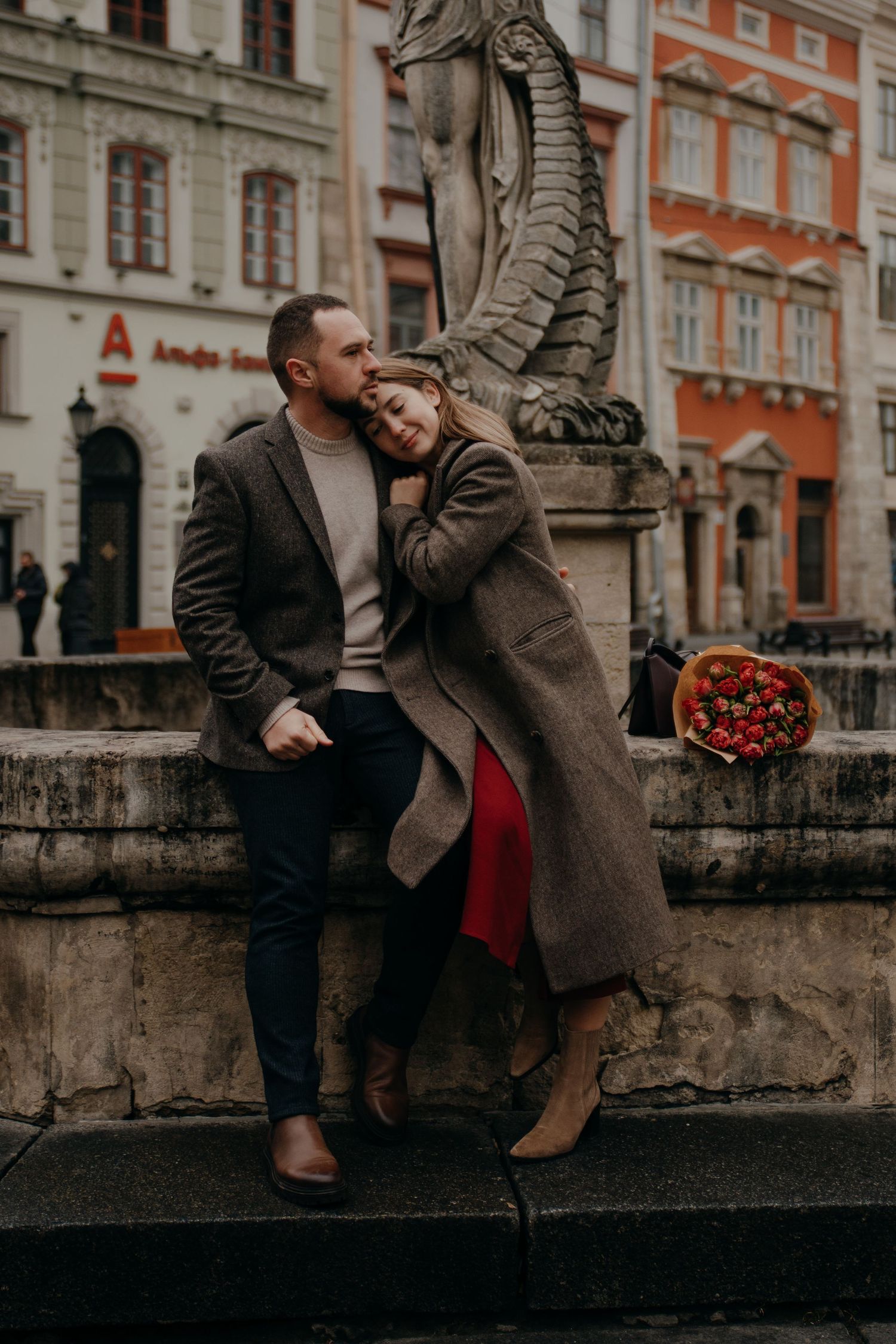 Romantic couple embracing by a stone dragon fountain in a European city square, woman in gray coat leaning on man.