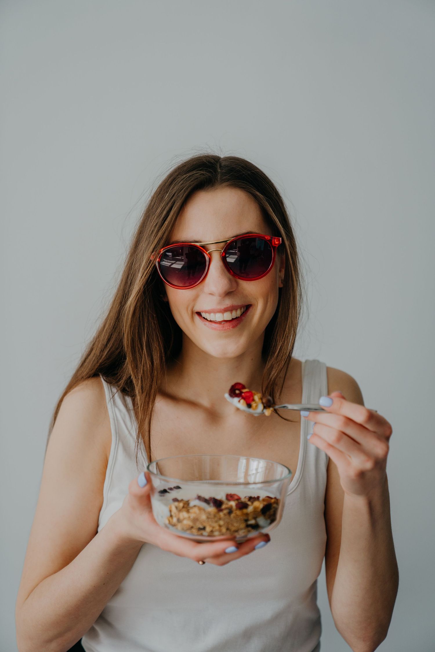 Smiling woman wearing red sunglasses eating a bowl of granola with berries on a light background.