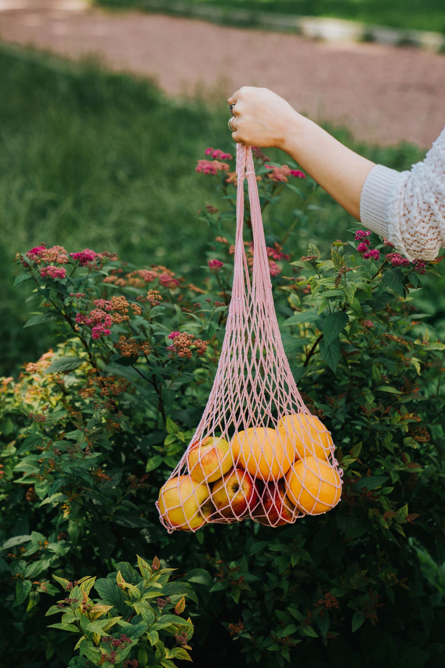 Hand holding a pink mesh reusable bag filled with fresh oranges and lemons outdoors near green flowering bushes.