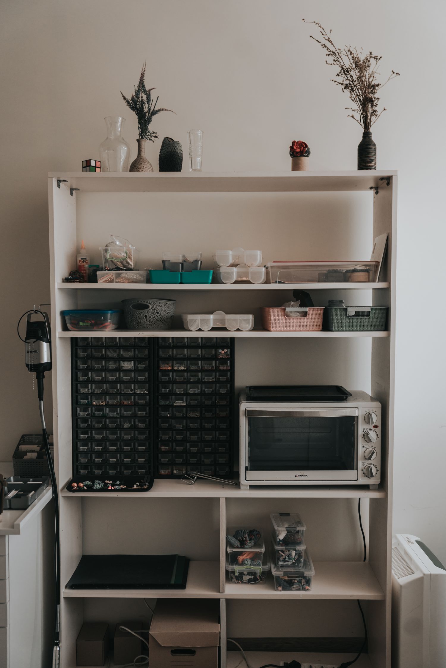 White shelving unit with organized craft supplies, a toaster oven, storage bins, and decorative plants on top.