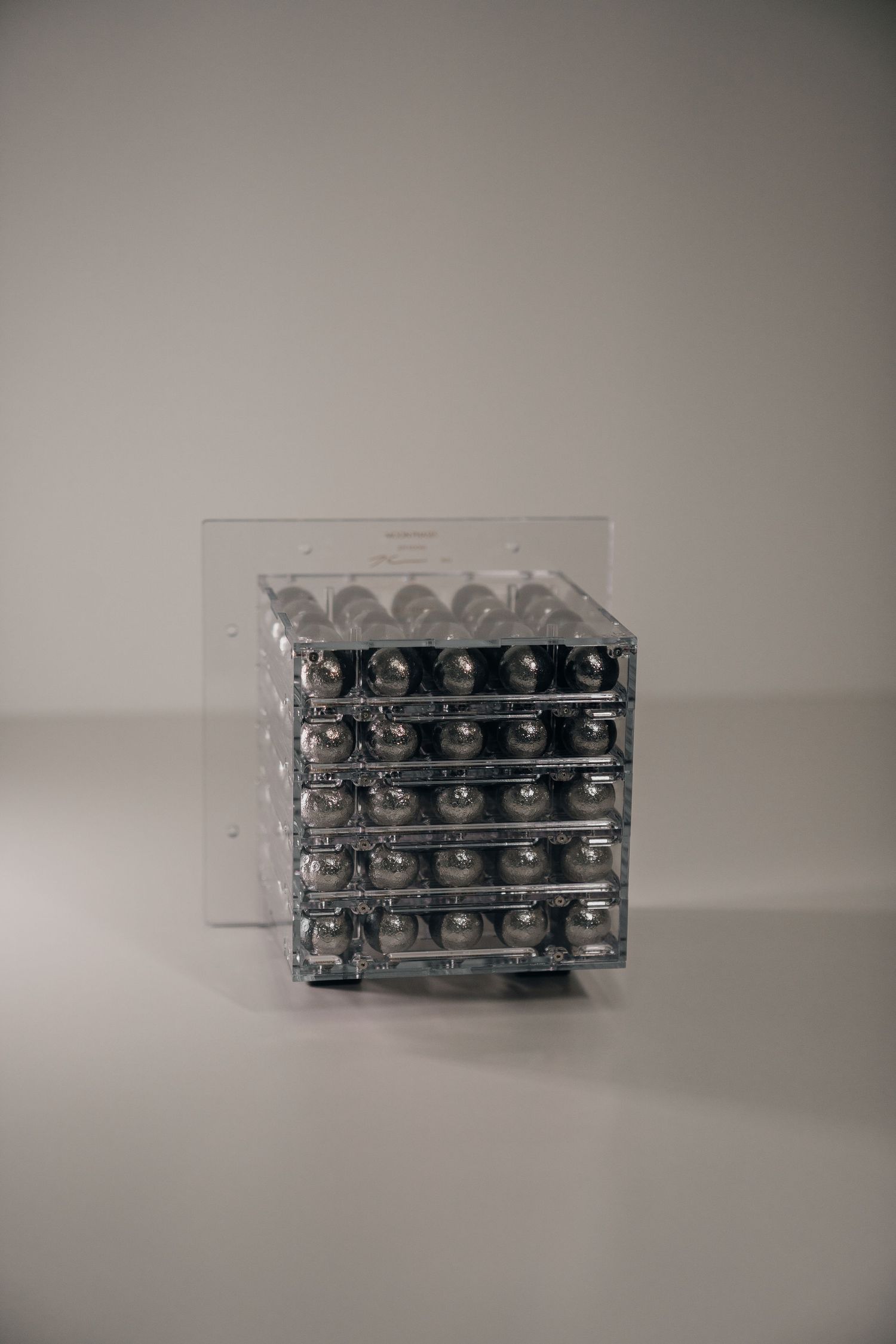 Clear acrylic cube display case filled with rows of shiny silver metal balls on a neutral background.